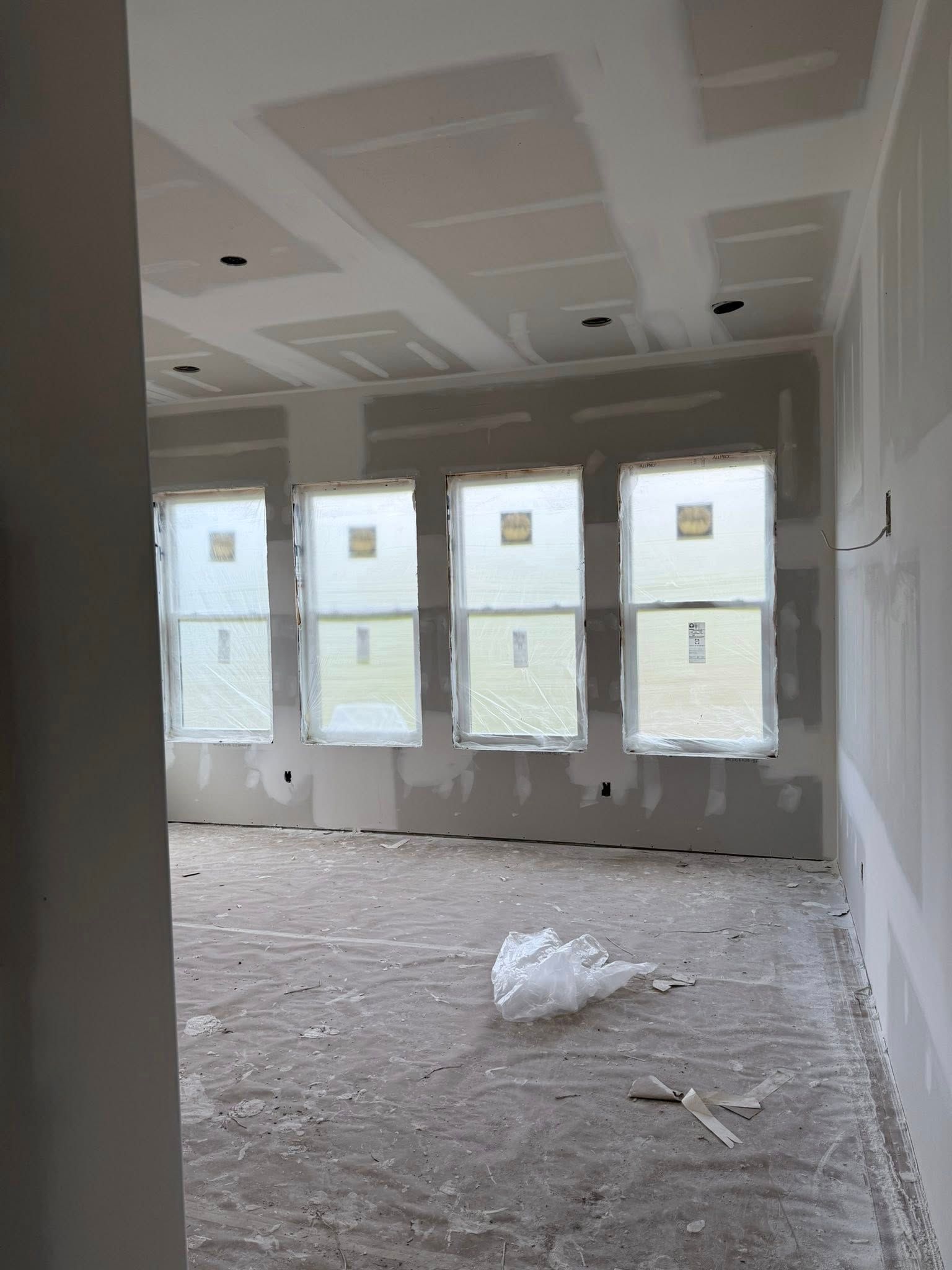 Interior of a room under construction with windows and drywall, covered in plastic and debris.