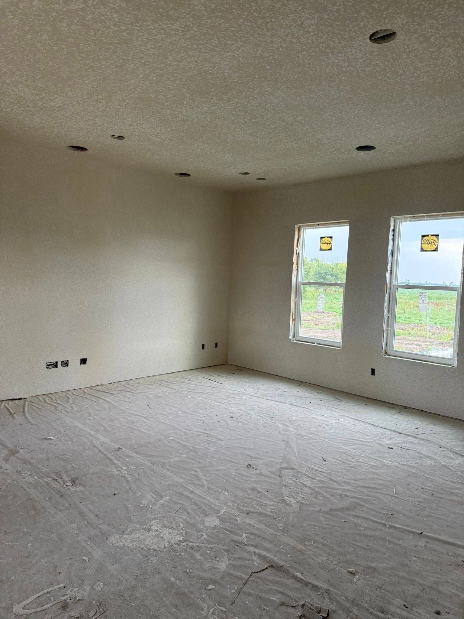 Empty room under construction; bare walls, unfinished floor, two windows, overhead light fixtures.