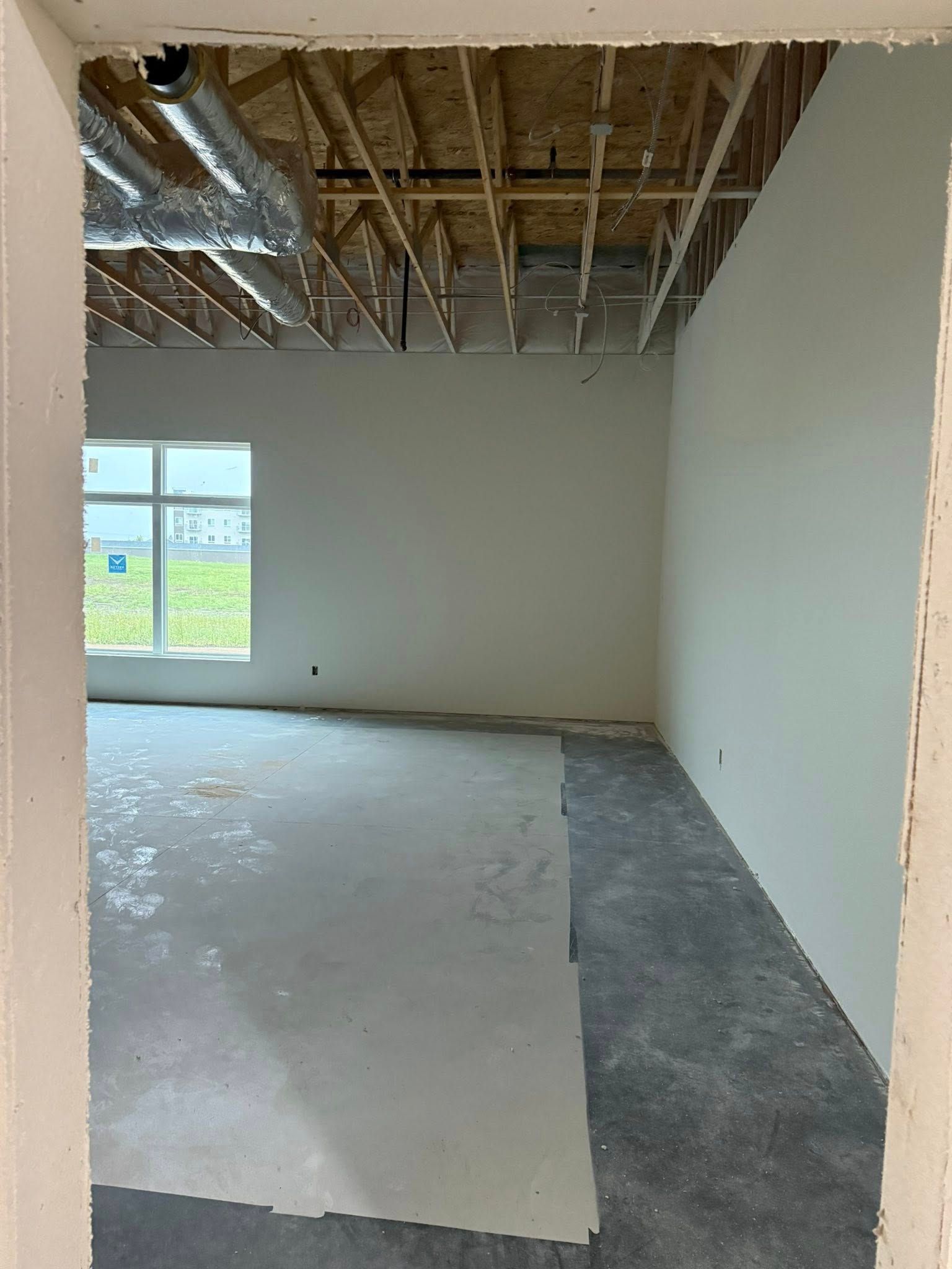 Interior view of unfinished room: white walls, gray floor, window, exposed ceiling beams, and a doorway.