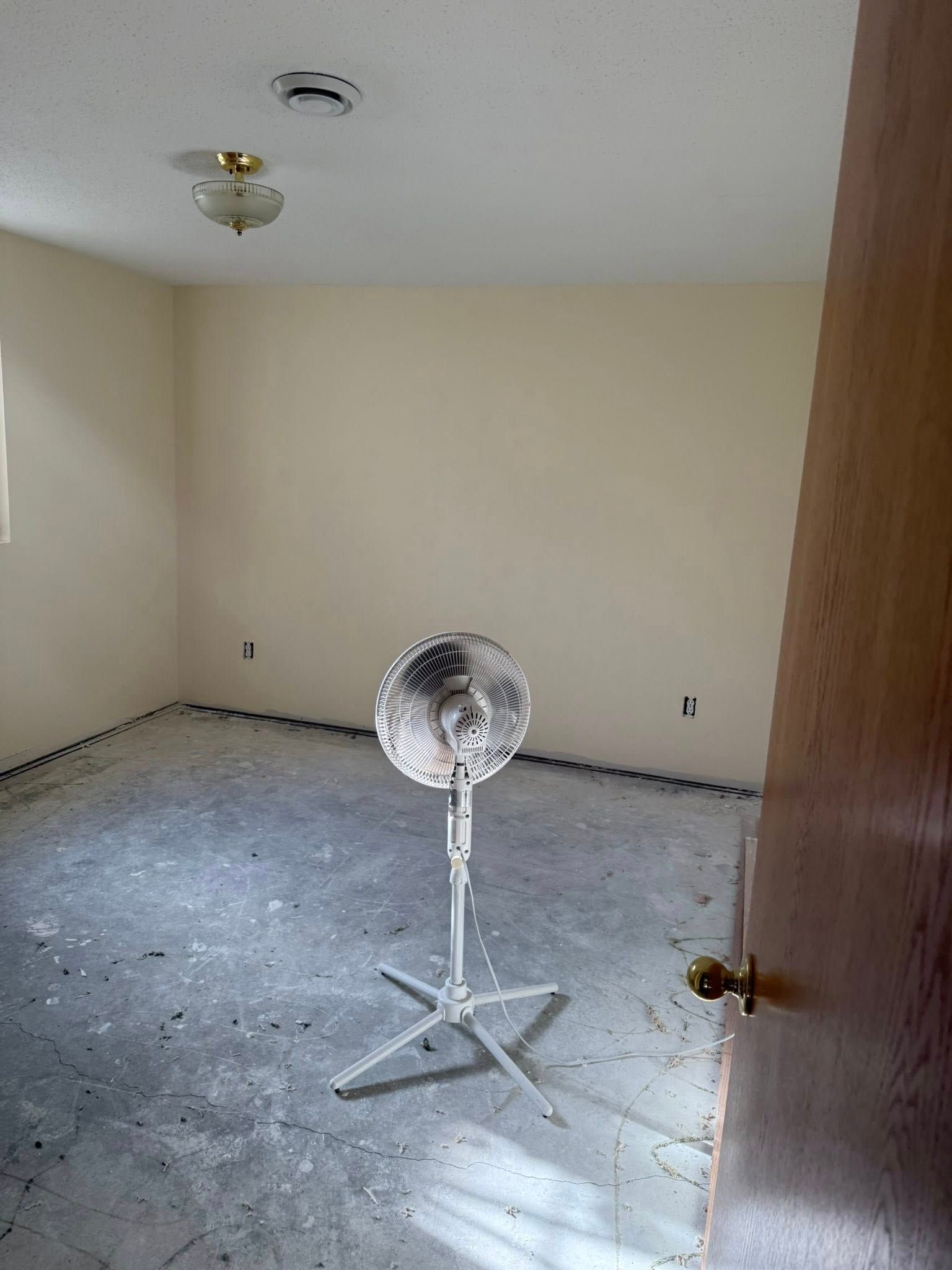 Room with tan walls, cement floor, ceiling light, smoke detector, and standing fan. Door on right.