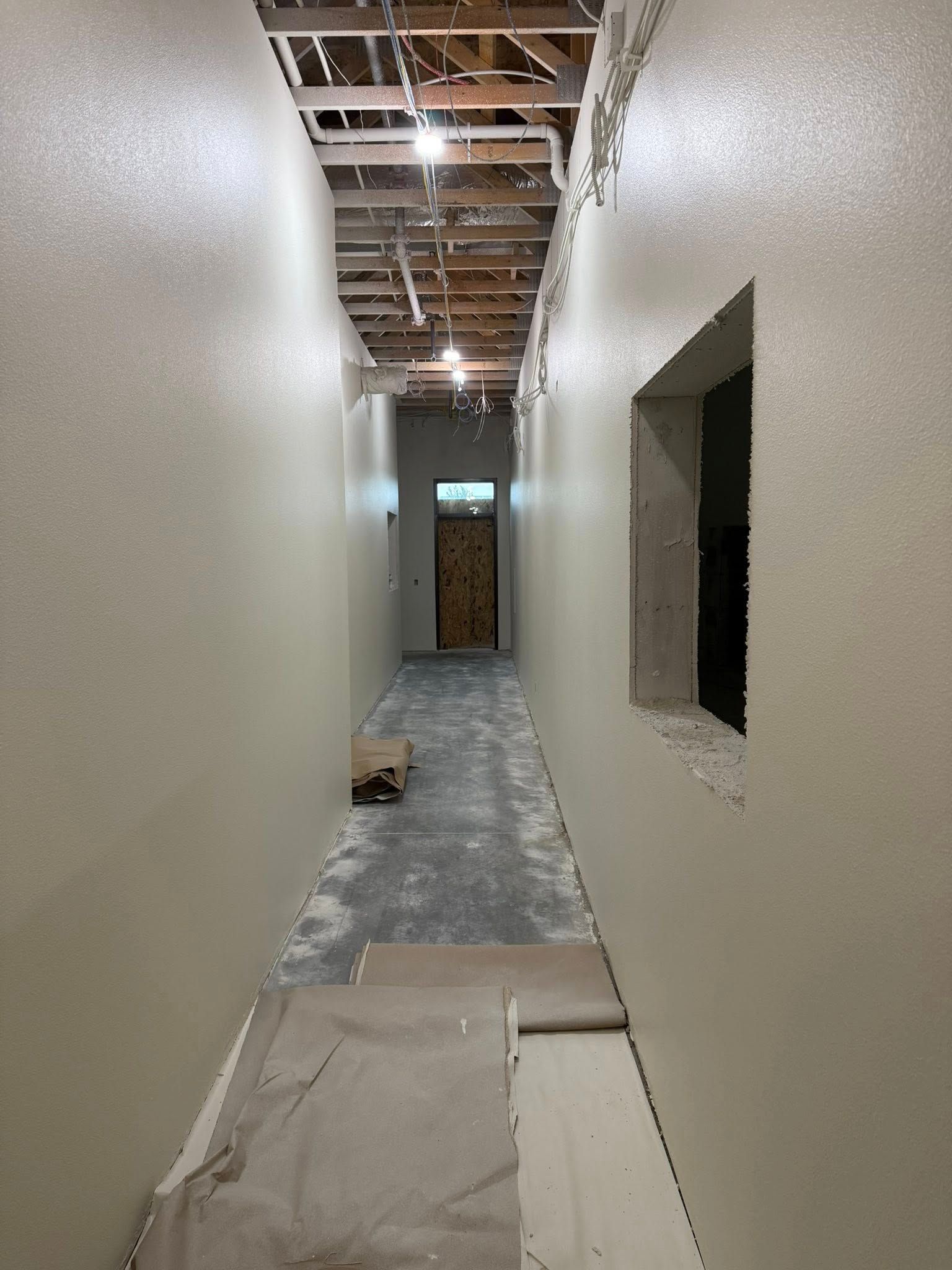Narrow hallway under construction, light-colored walls, unfinished ceiling, window on right, doorway at end.