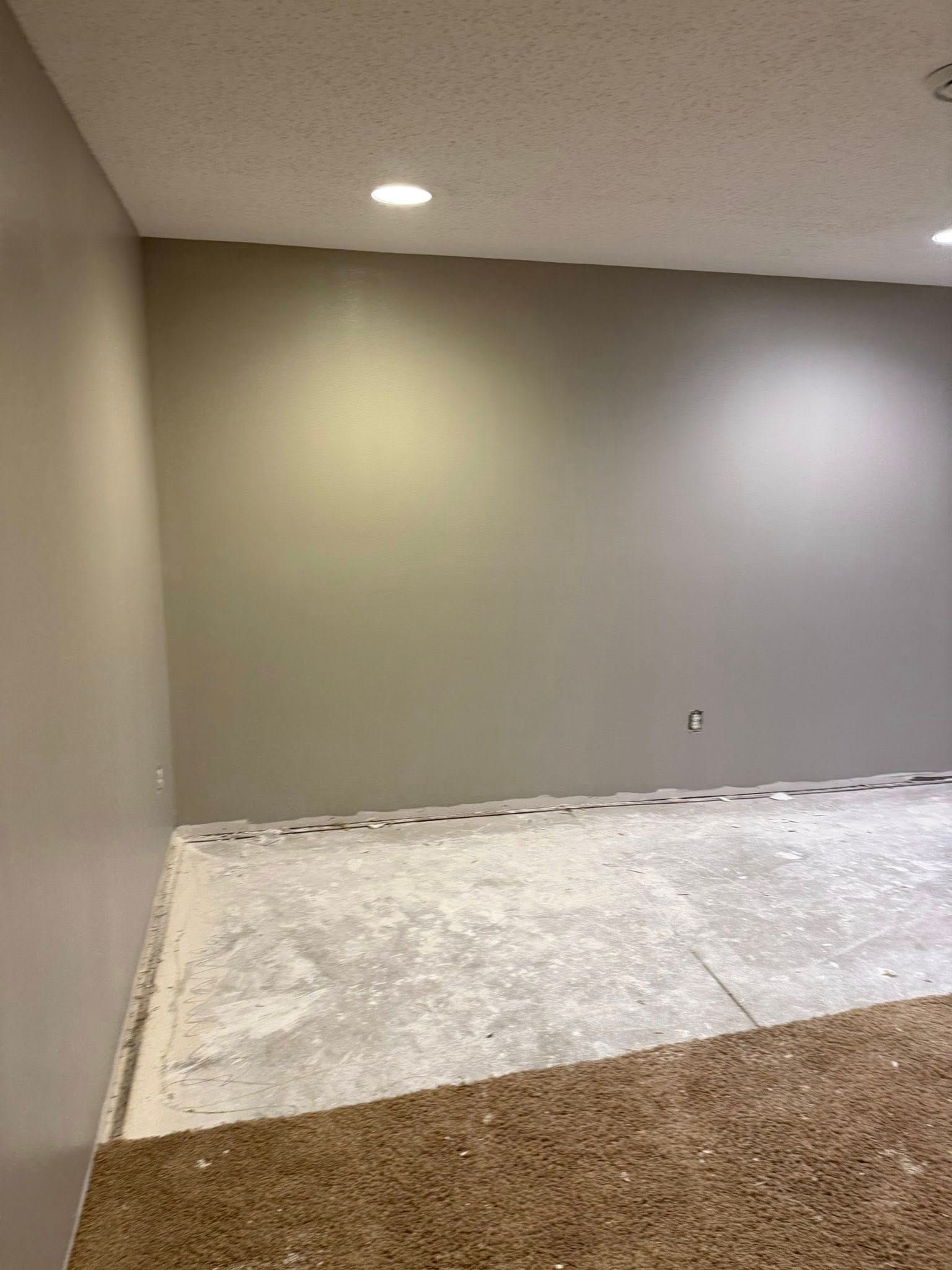 Empty room with neutral walls, exposed concrete floor, and carpeted section. Recessed lighting illuminates.
