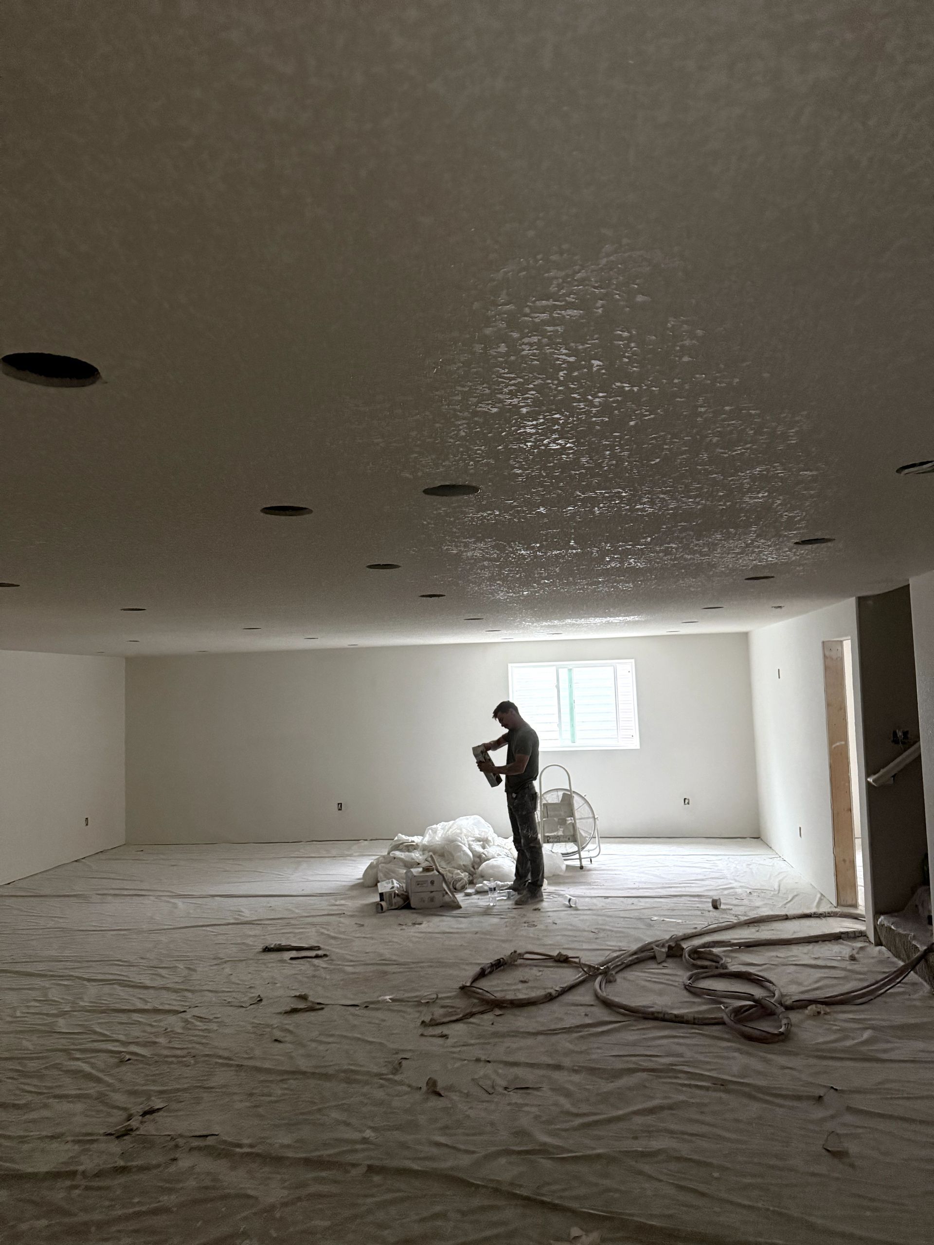 A basement room with gray walls and a brown carpet. A fan sits on the carpet near exposed flooring.