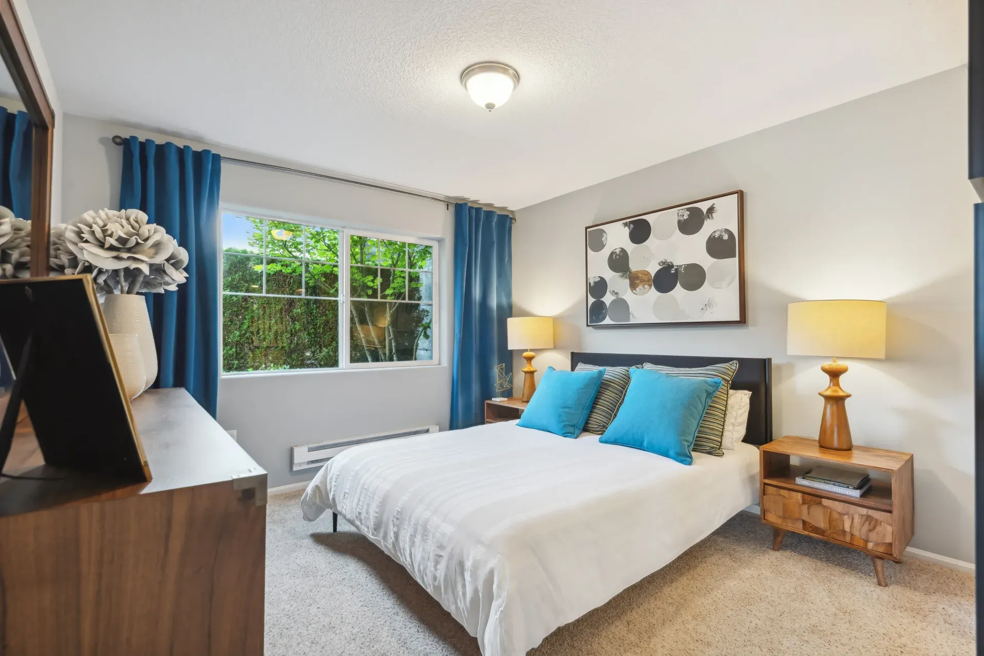 Bedroom with bed, blue curtains, and artwork. Wooden nightstands with lamps flank the bed.