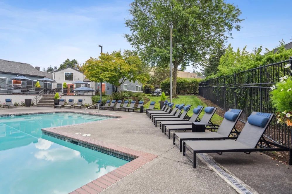 Swimming pool with lounge chairs, trees, and buildings under a blue sky.