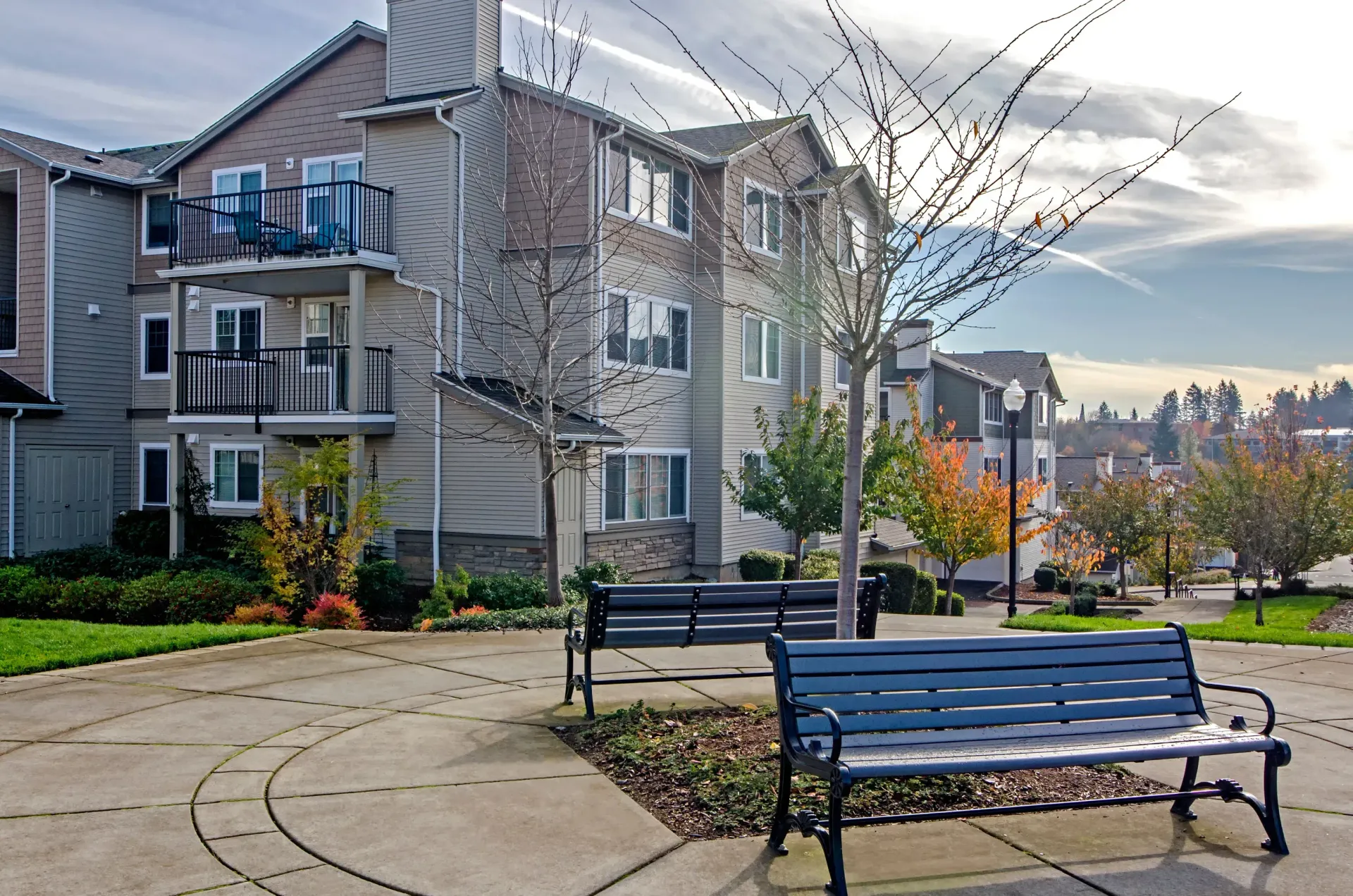Apartment building with benches in a paved courtyard. Trees with changing leaves and blue sky.