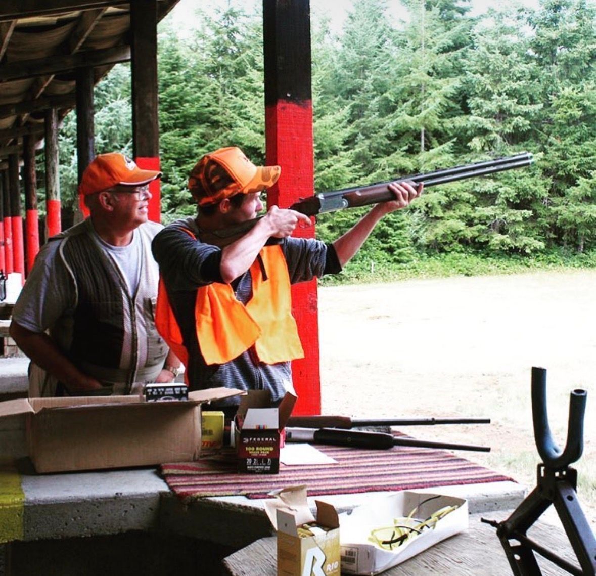 Two people at a shooting range aiming a shotgun under a covered outdoor shelter.