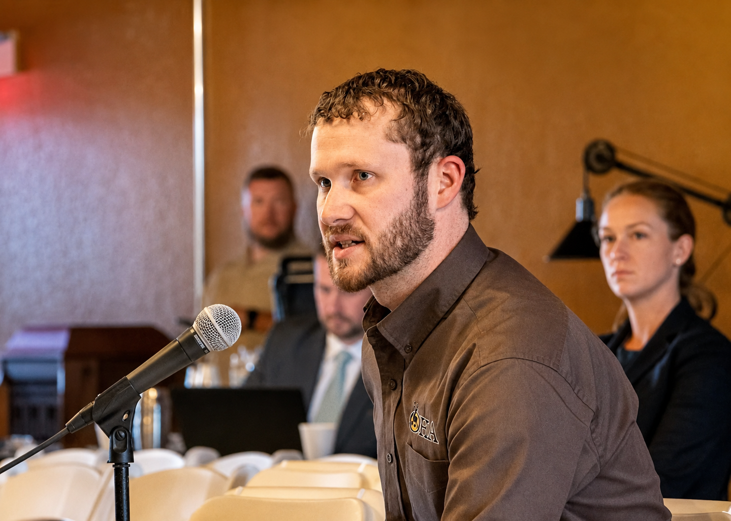 Man speaking into a microphone at a conference table, with a blurred audience behind him.