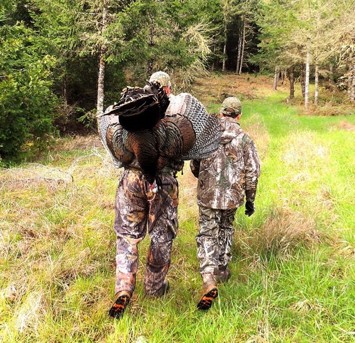 Two camouflaged hikers walking through a grassy forest trail, one carrying a large netted bundle.