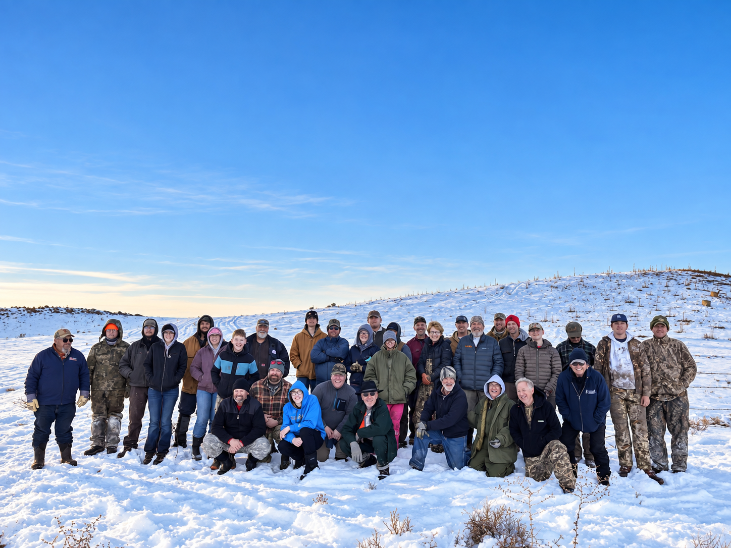 Group of people standing and kneeling in snowy field under a bright blue sky, wearing winter gear and camouflage.