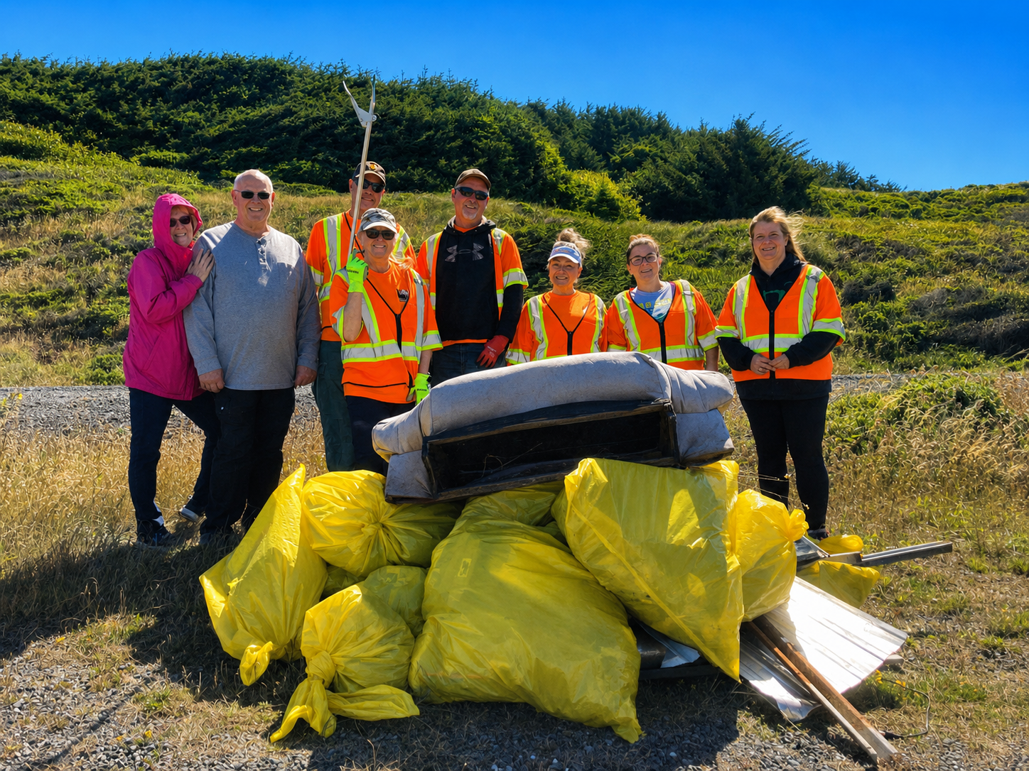 Cleanup crew posing with trash bags and litter picker in a sunny grassy field near a hedgerow.