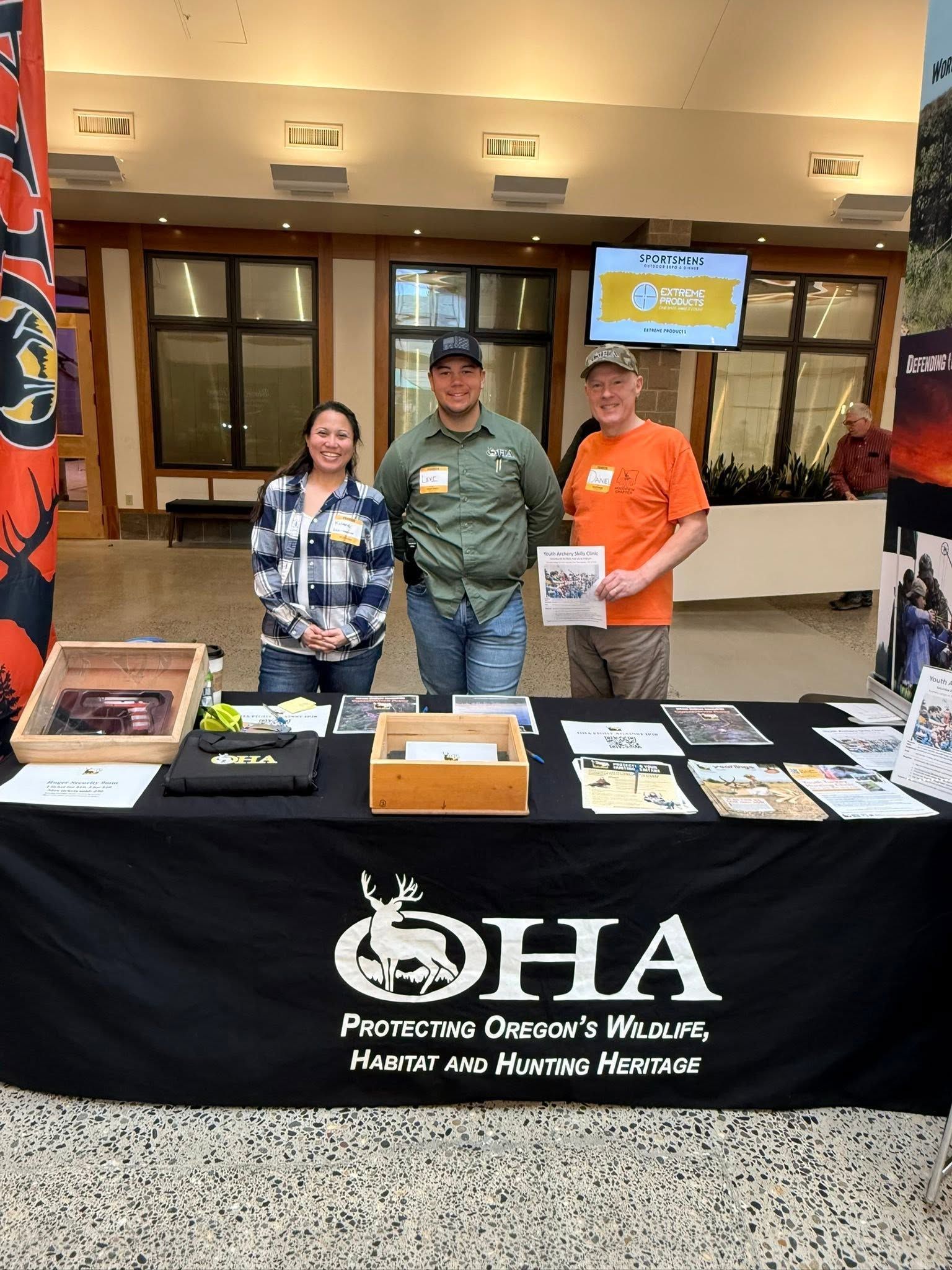 Three people stand behind a resource table with a black OHIA tablecloth at an indoor event.