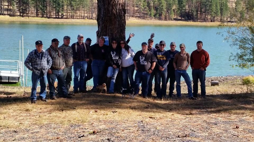 Group of people posing outdoors beside a lake, with trees and a dock in the background.