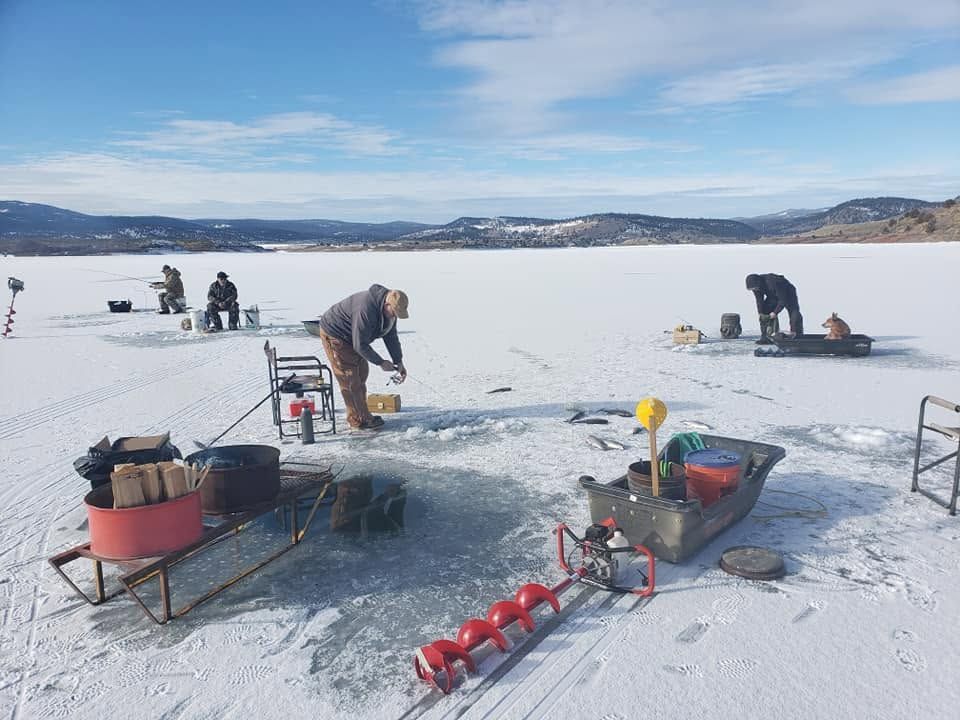 People ice fishing on a frozen lake with drills, sleds, and gear under a cloudy blue sky