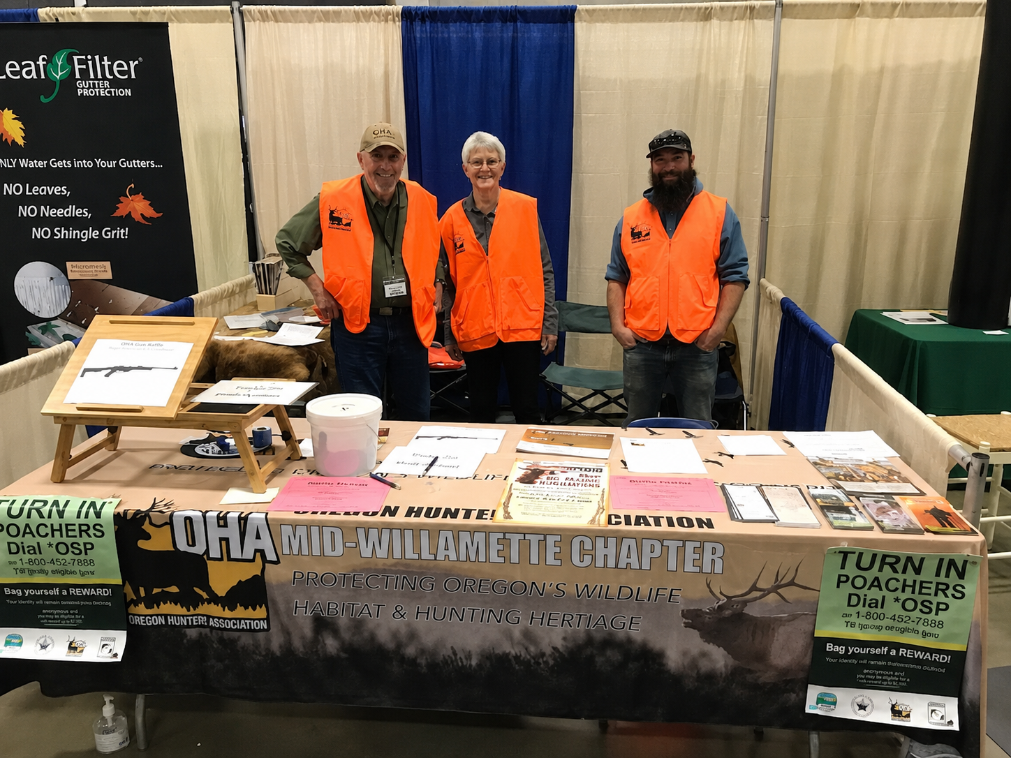 Three volunteers in orange safety vests stand behind a resource booth with pamphlets and signs.