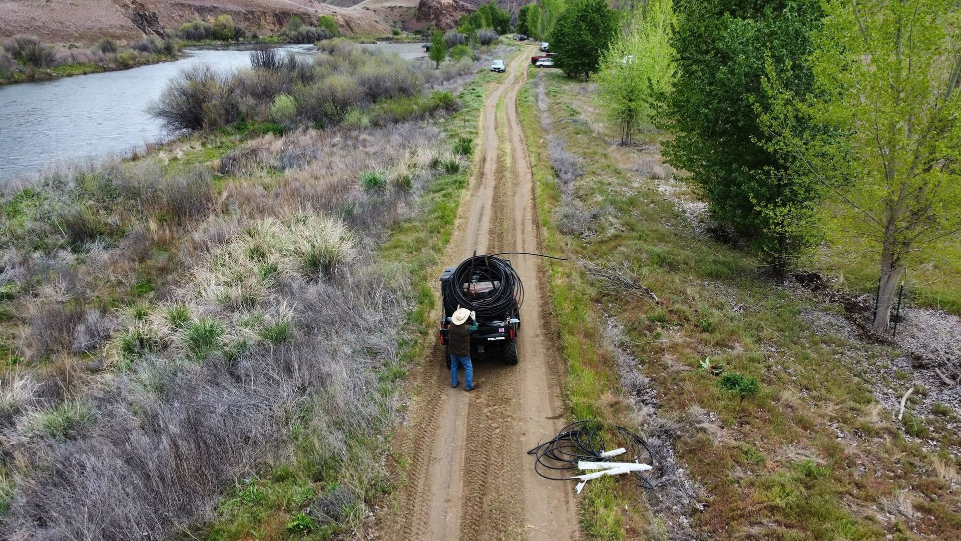 Black ATV on a dirt trail beside a river, with one person standing nearby in a rural landscape.
