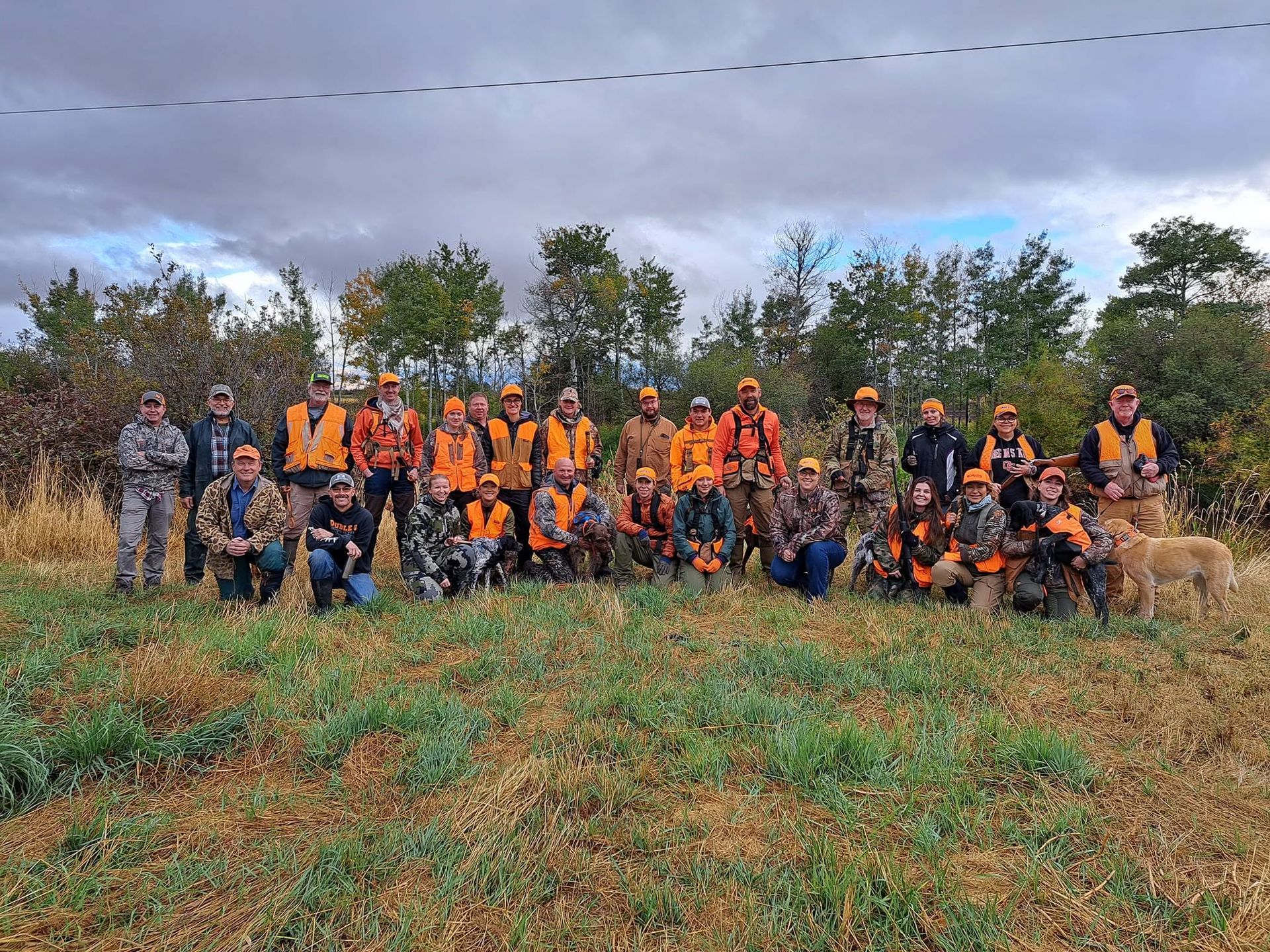 Group in orange safety vests posing outdoors in a grassy field with trees and cloudy sky