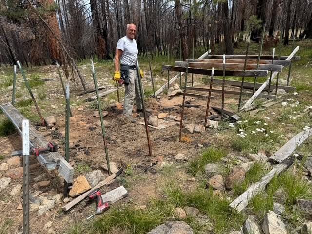 Man standing in a rocky backyard beside a partially built metal framework foundation.