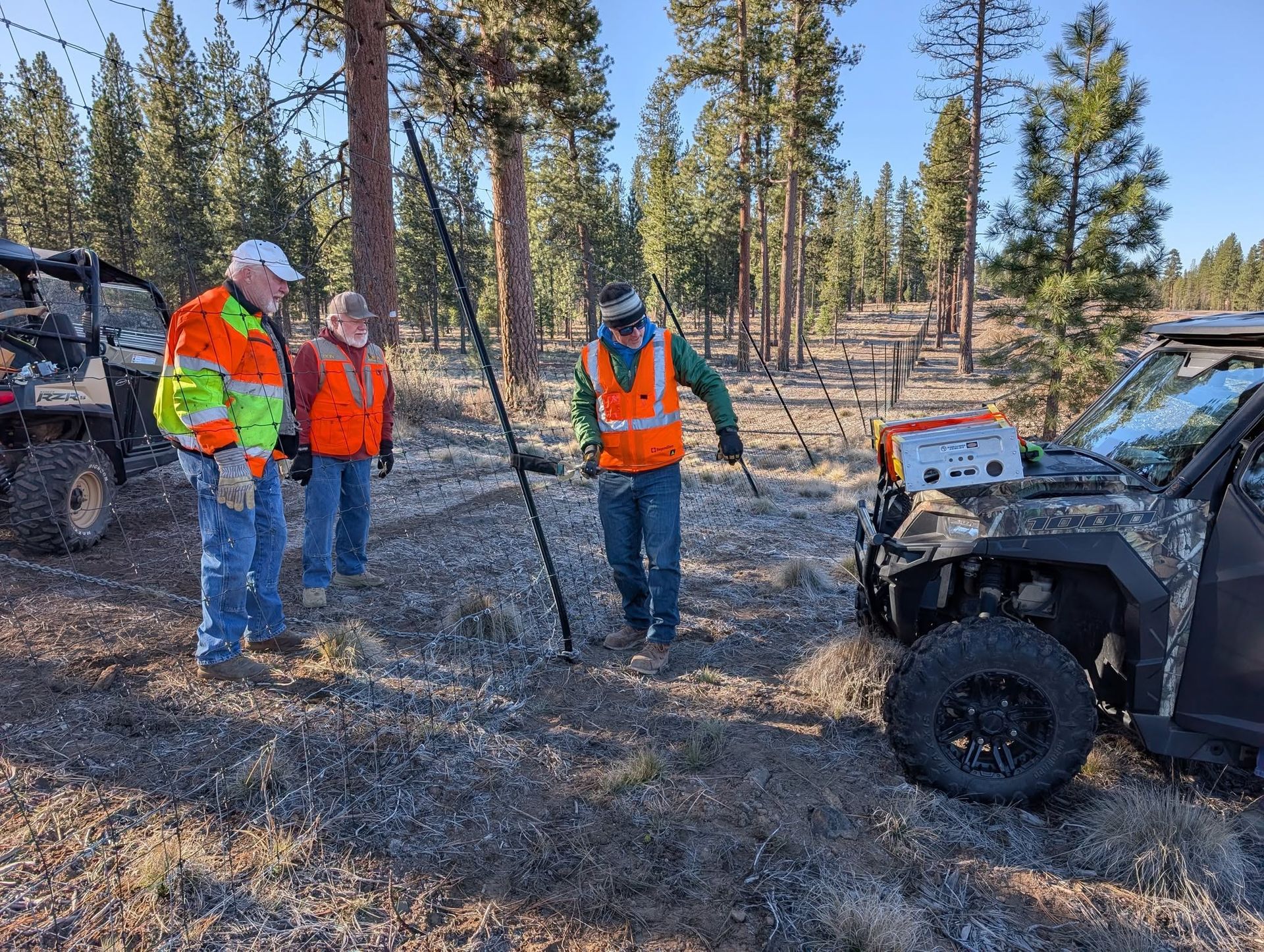 Workers in orange safety vests inspect a utility vehicle in a forest clearing.