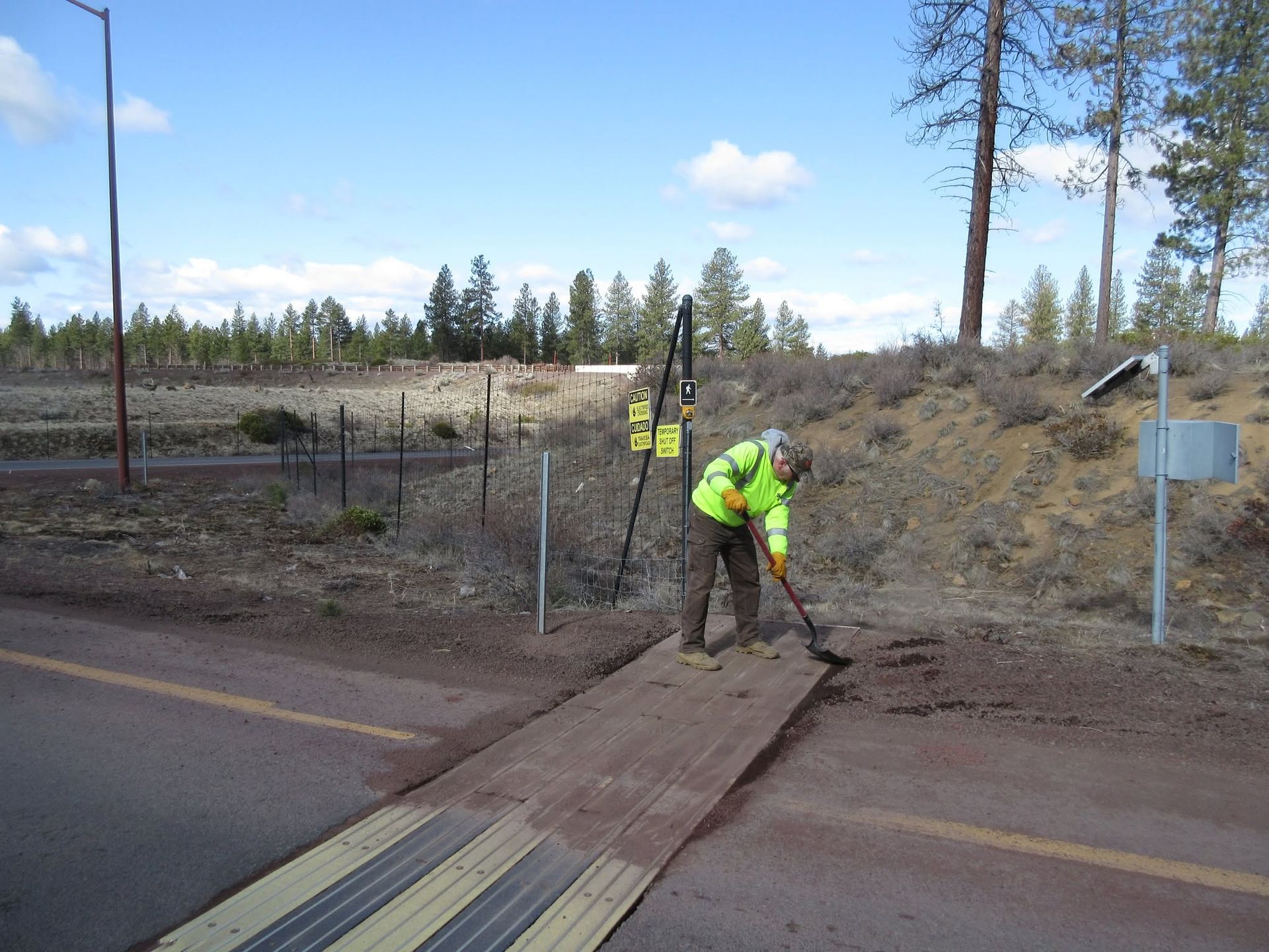 Worker in a neon jacket shovels dirt beside a road and fence in a rural area.