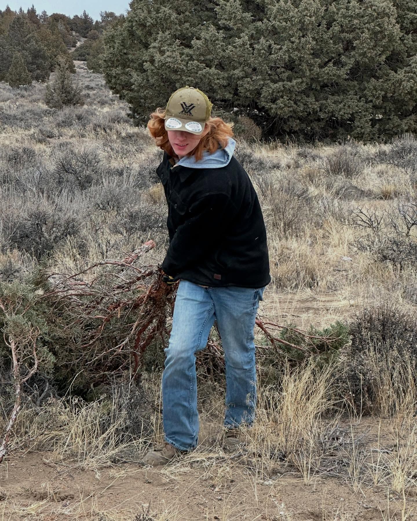 Person in a cap and black jacket standing in a grassy field holding a bundle of branches.