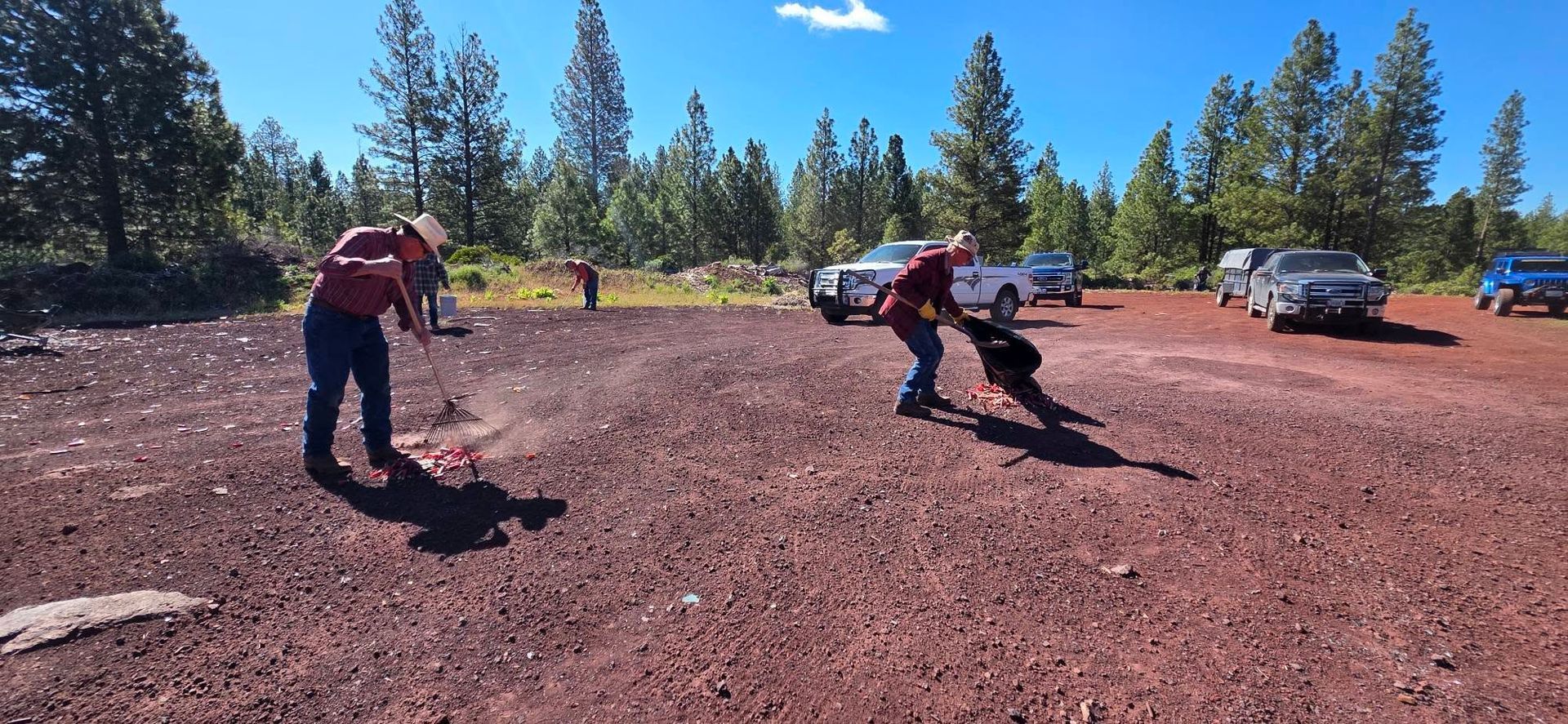 Two people working in a red dirt clearing with trees and parked cars in the background.
