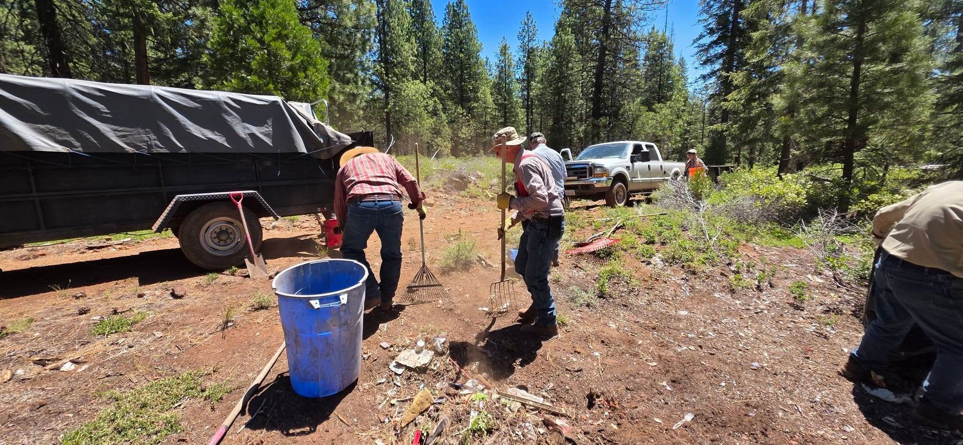 People working outdoors beside a trailer in a forest clearing, with a blue barrel and a white truck.