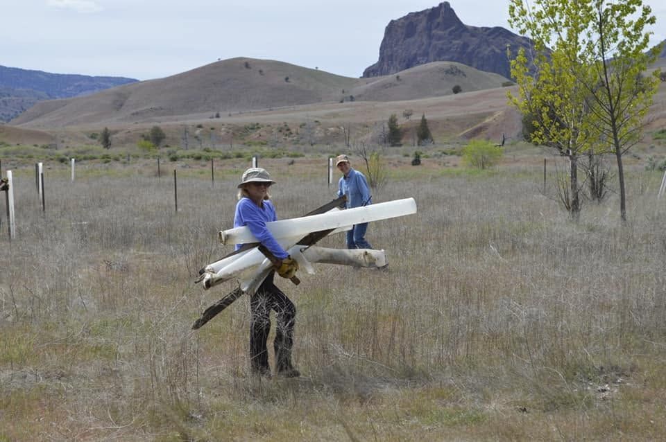 Two people in a grassy field carry a large white model aircraft near rocky hills.