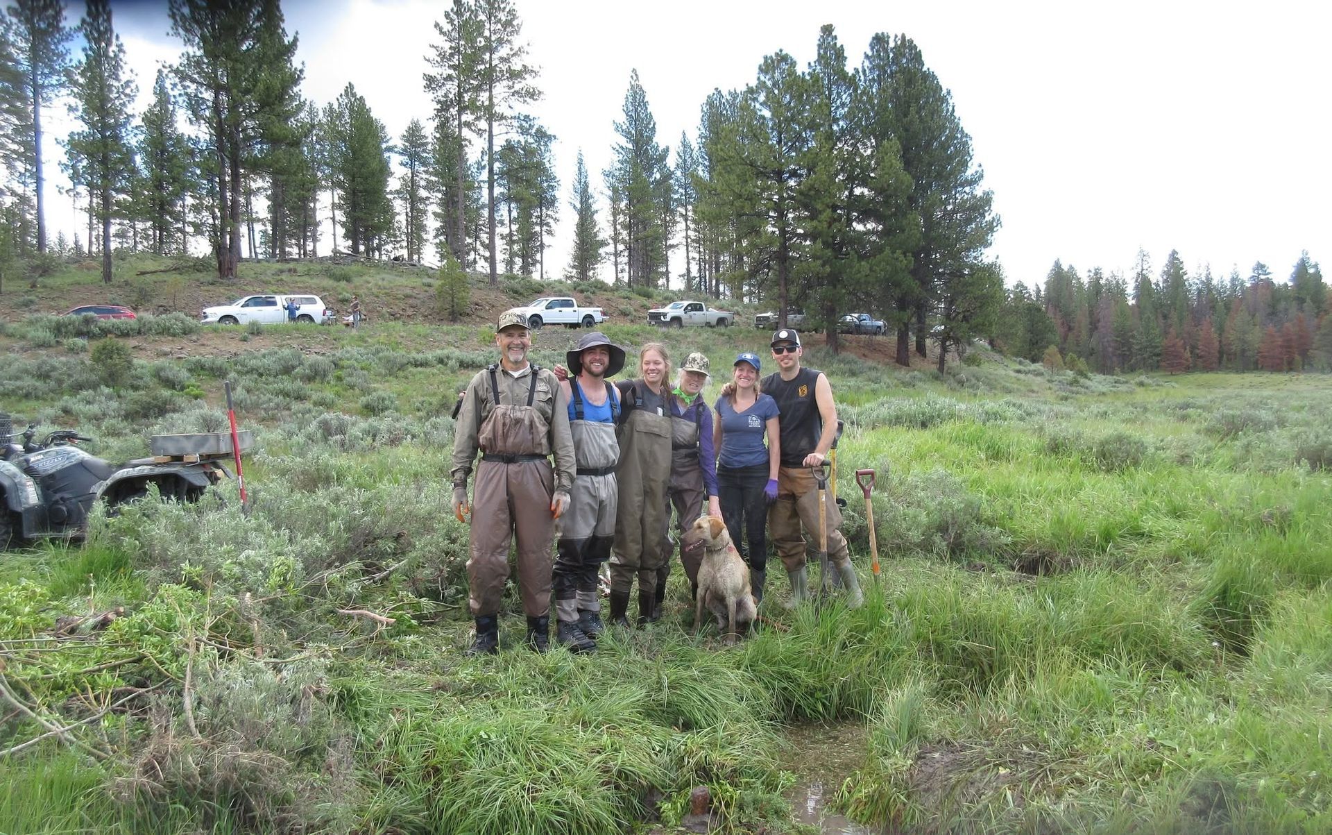 Five muddy hikers pose together in a grassy forest clearing with trees in the background.