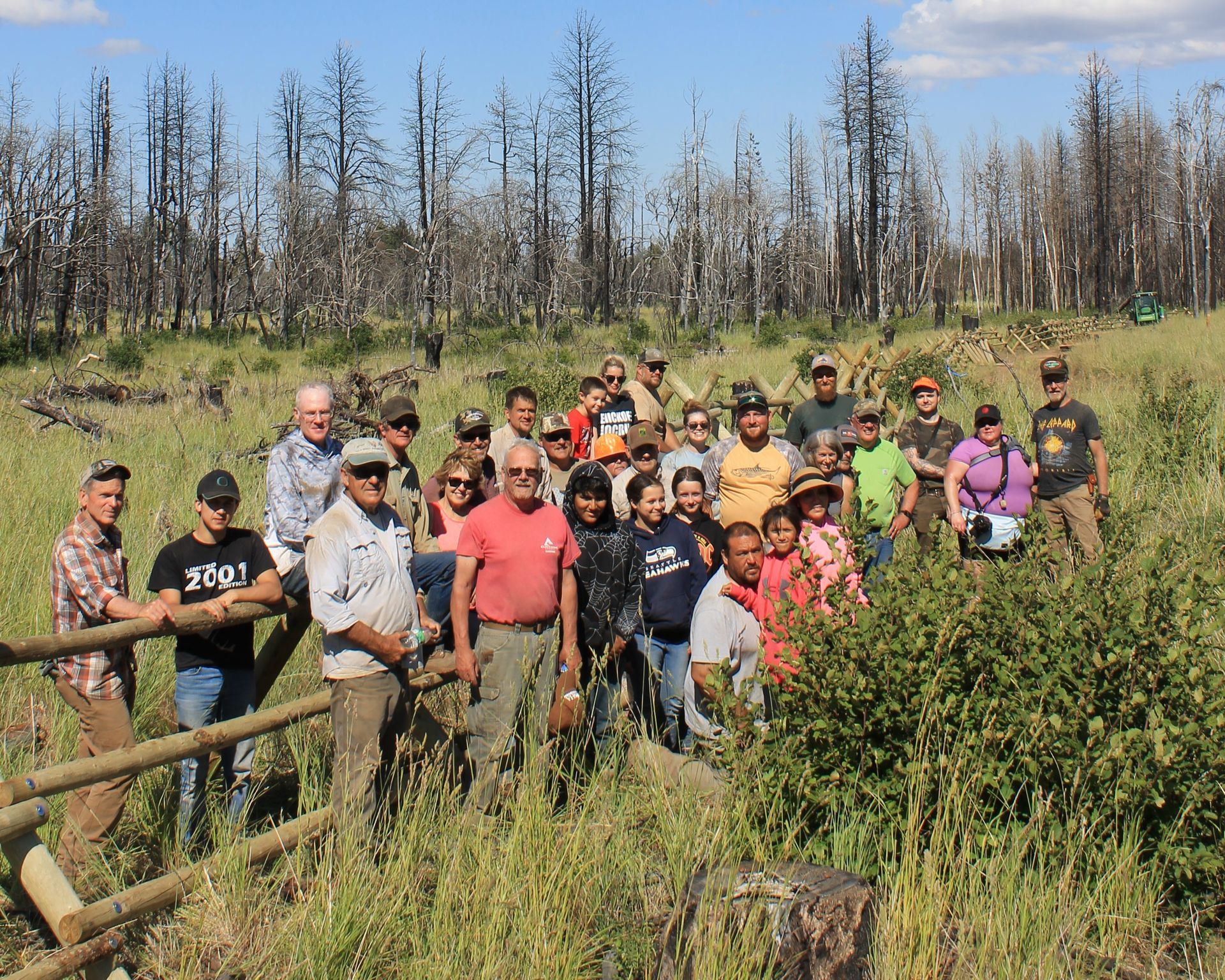 Group of hikers posing on a wooden bridge in a grassy area with charred trees under a blue sky