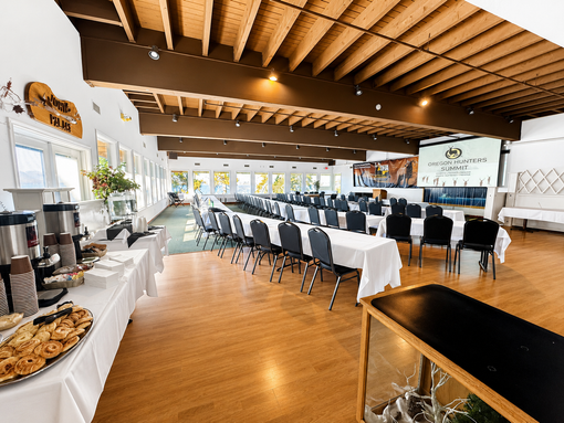 Modern banquet hall with long tables, white tablecloths, black chairs, and wood floors under a slatted ceiling
