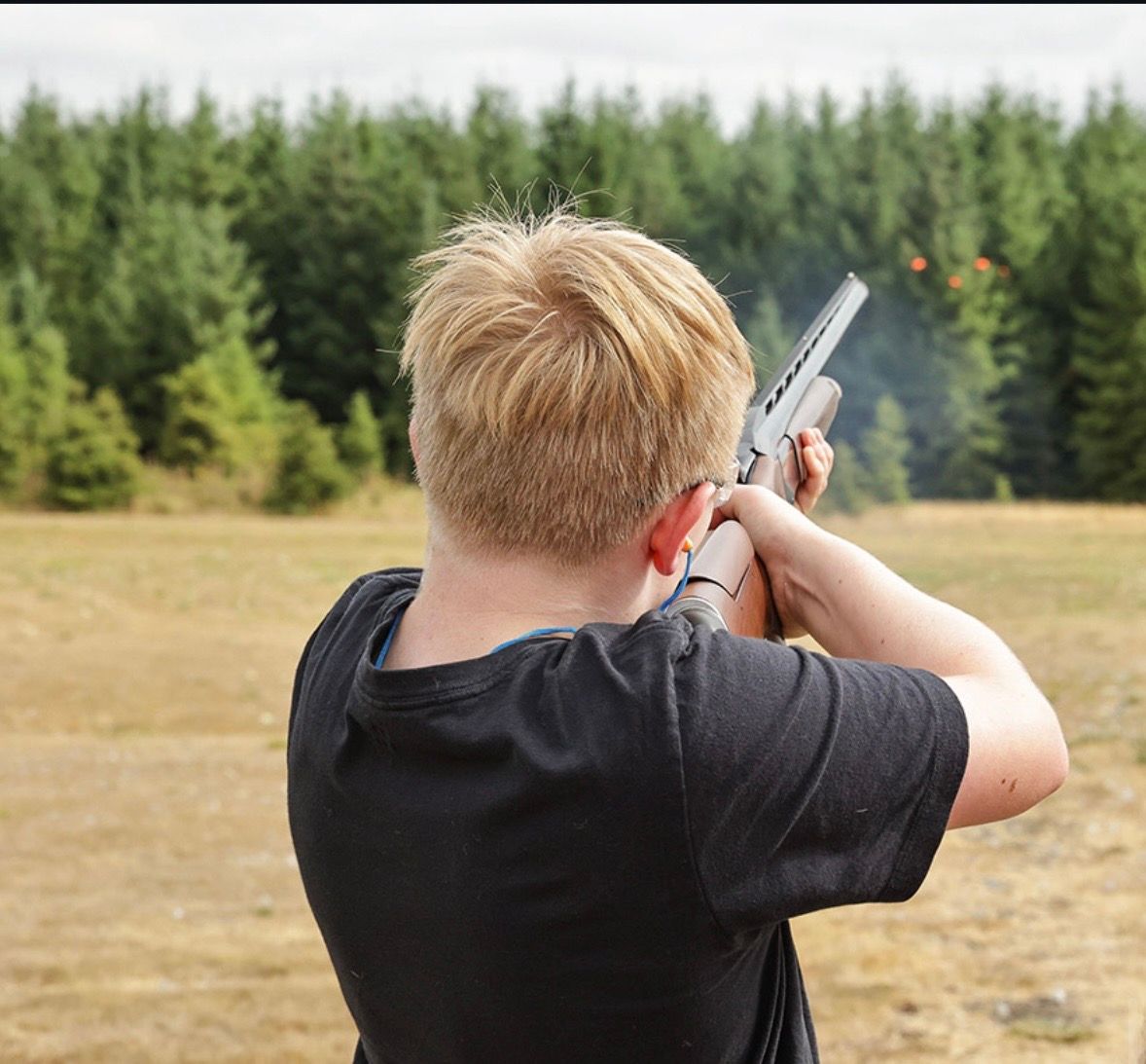 Child aiming a silver rifle at a target in a grassy field with trees in the background