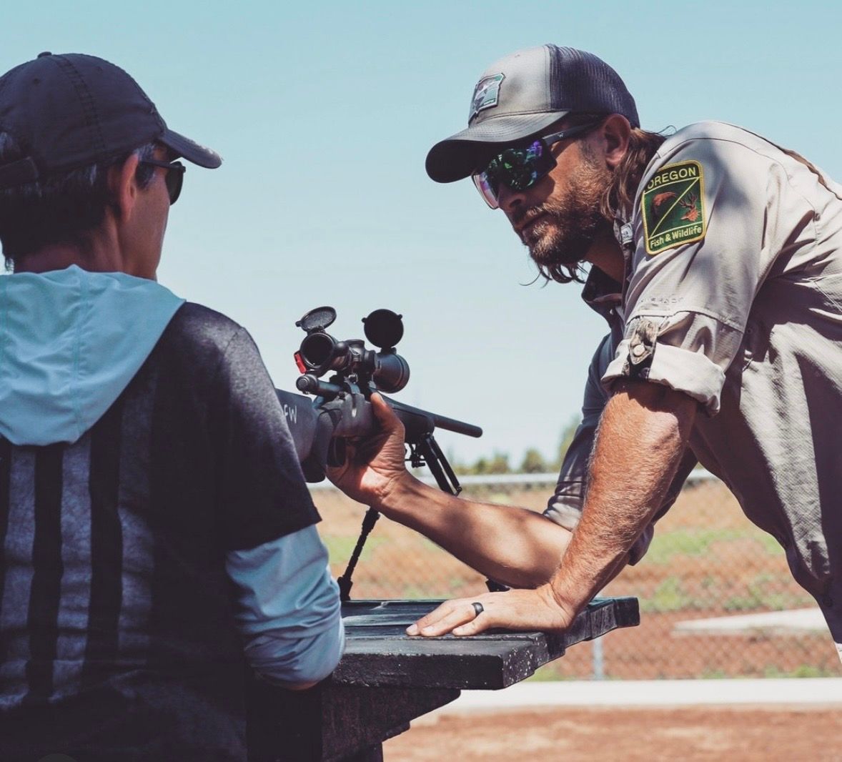 Instructor demonstrating a scoped rifle to a trainee at an outdoor shooting range