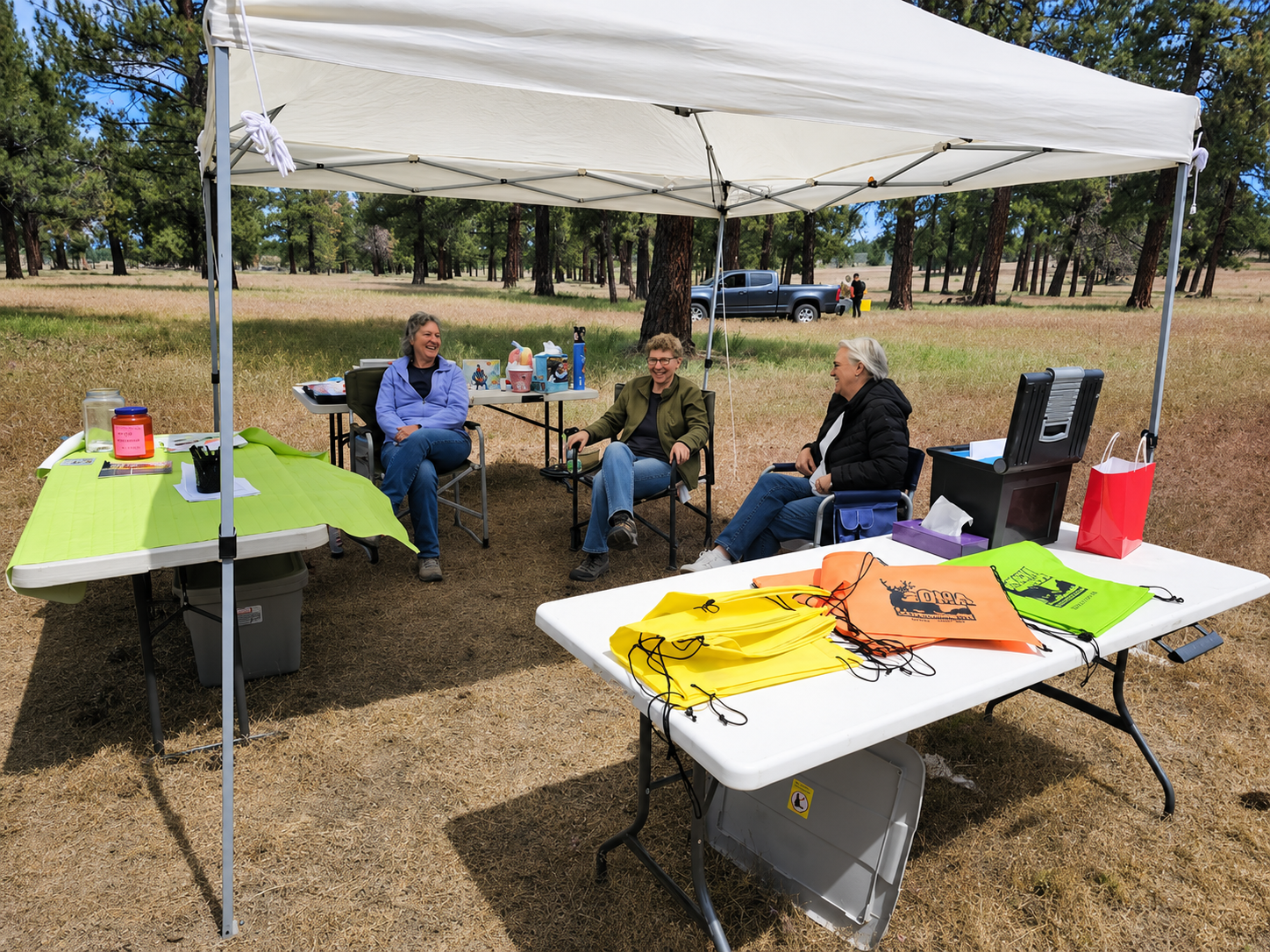 People seated under a white canopy at an outdoor table with craft materials and colorful papers.