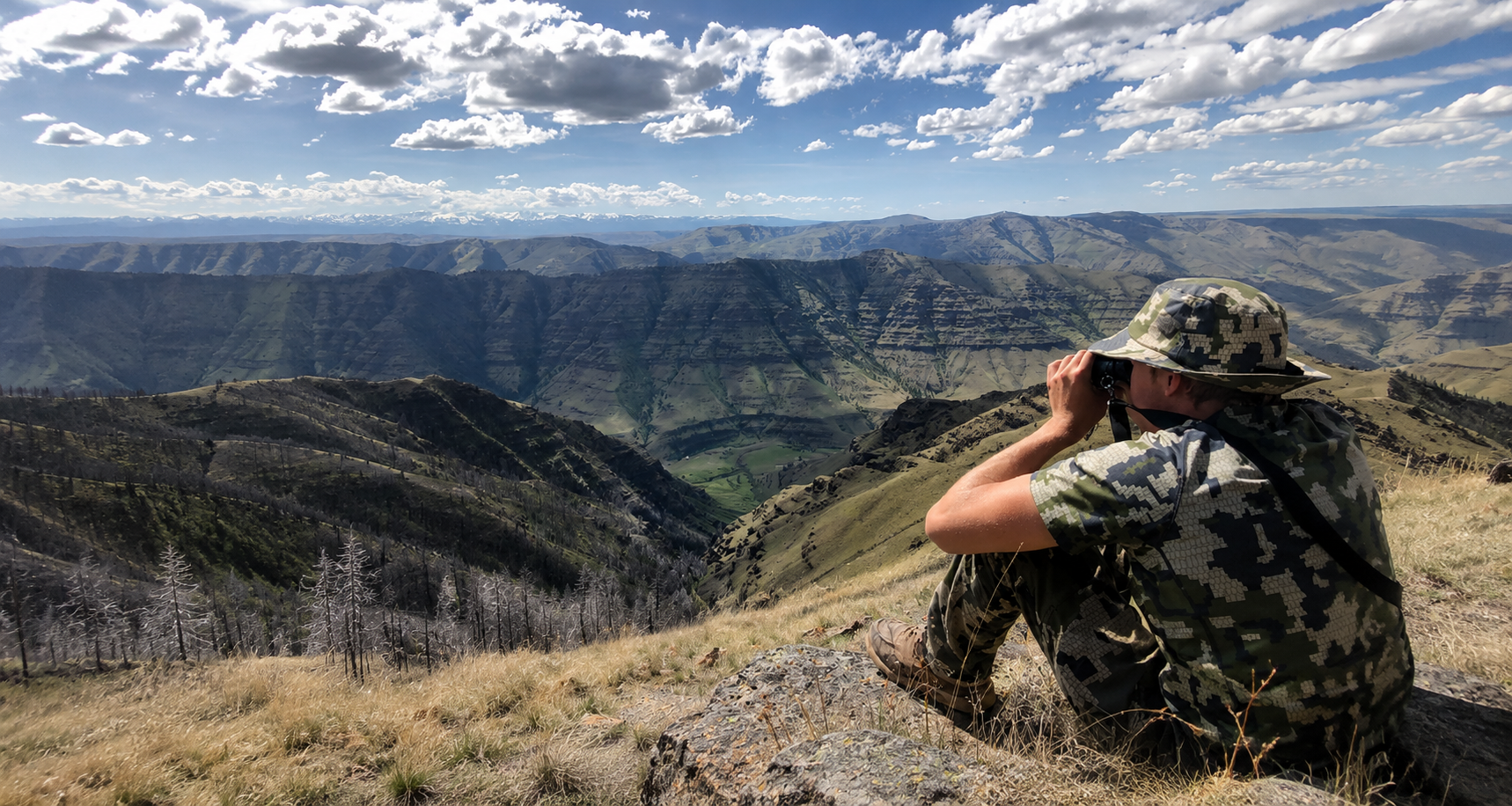 Person in camouflage sits on a rocky ridge, looking over a wide mountain valley under a cloudy sky