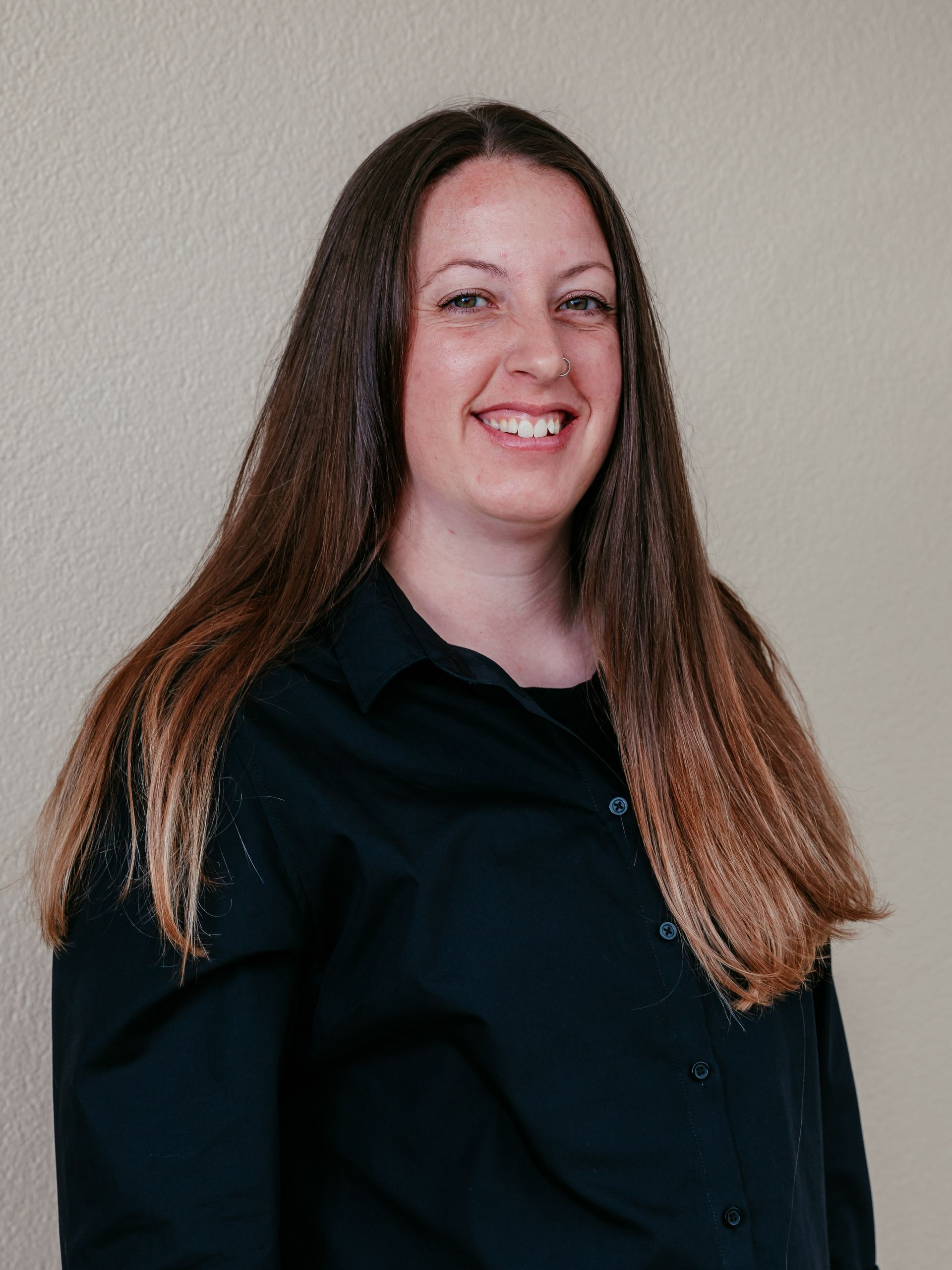 Smiling woman with long brown hair in a black shirt against a light gray wall