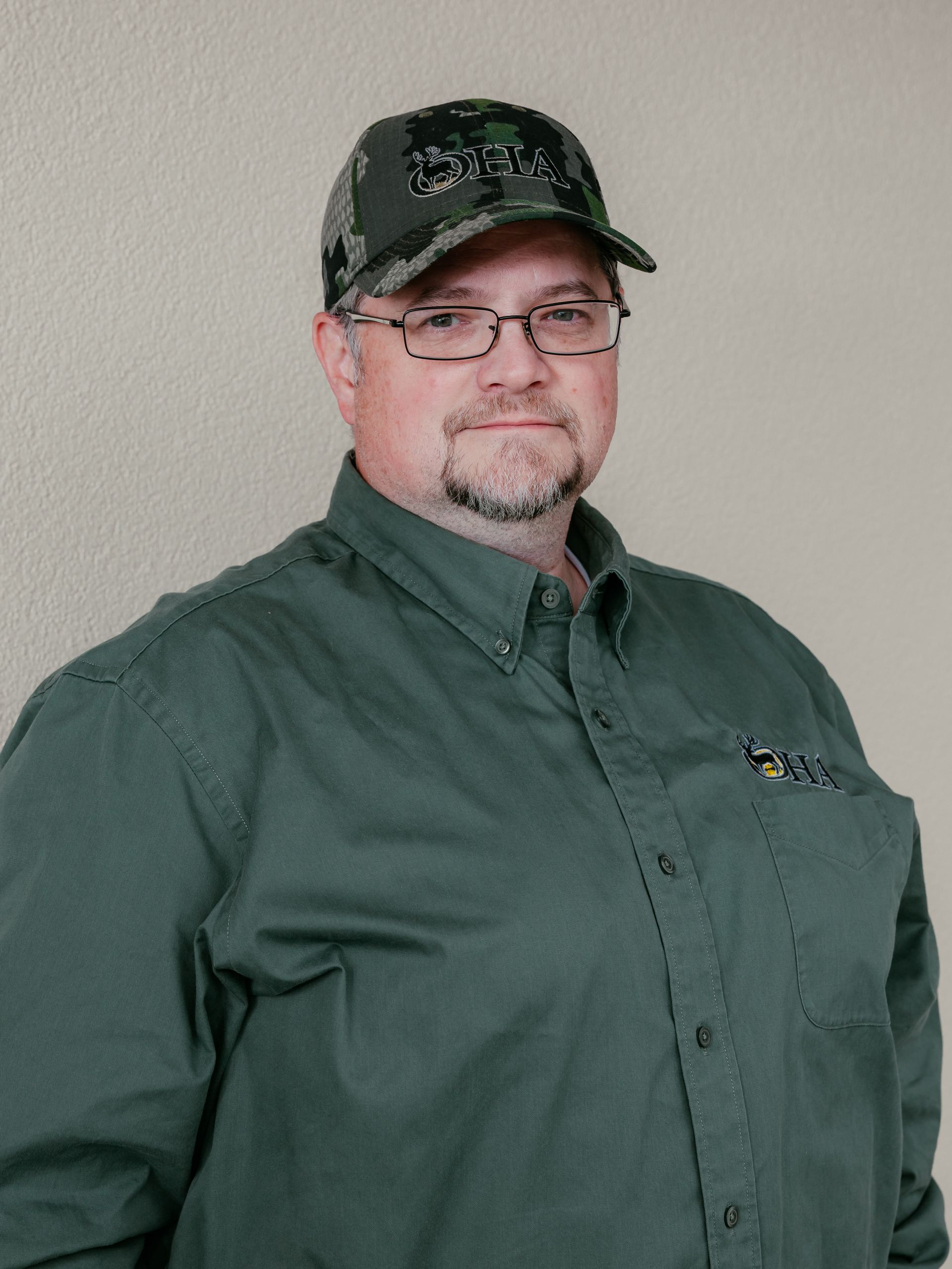 Person in a green button-up shirt and camo cap standing against a plain wall.