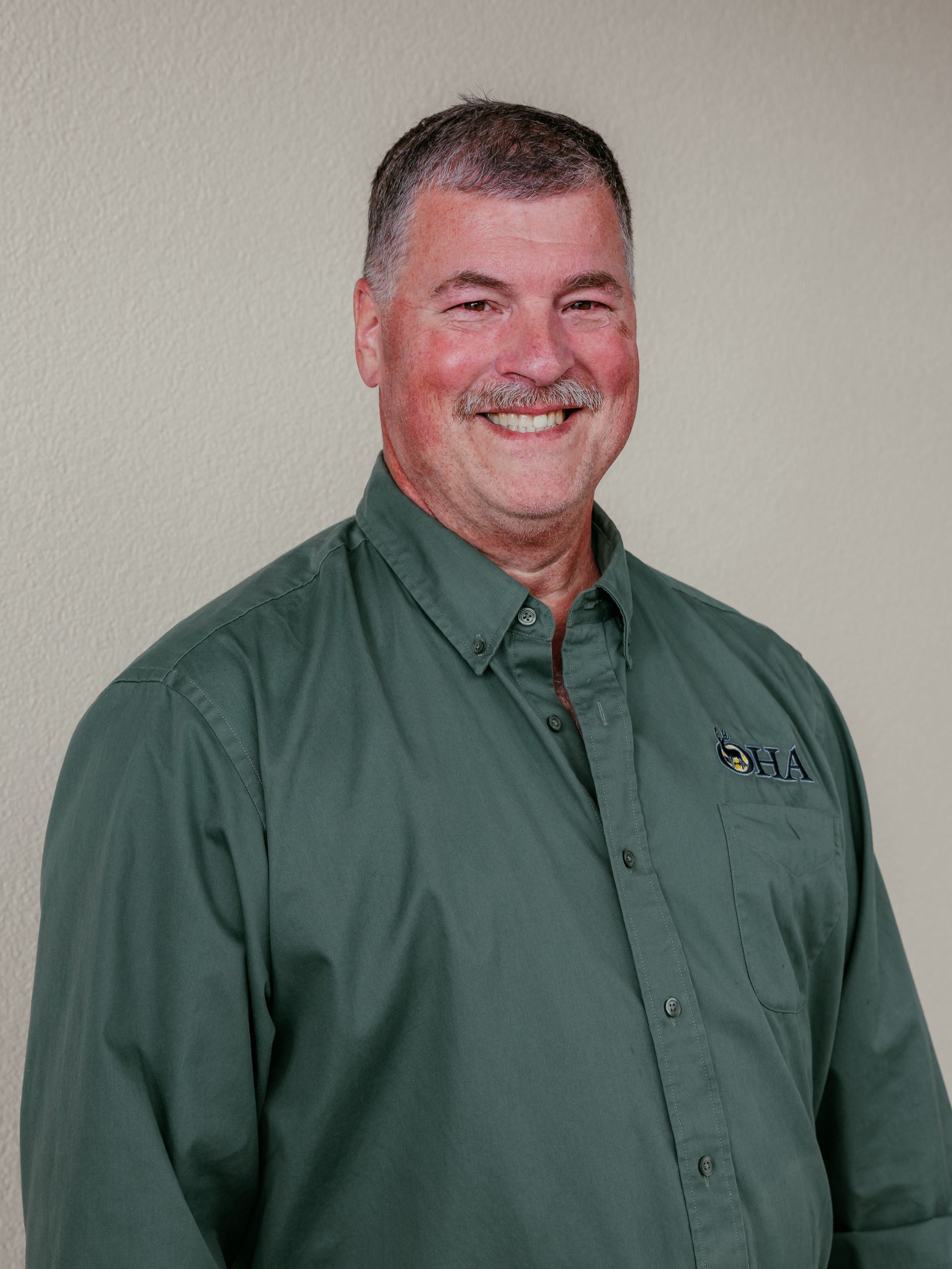 Smiling man in a green button-up shirt standing against a plain beige wall