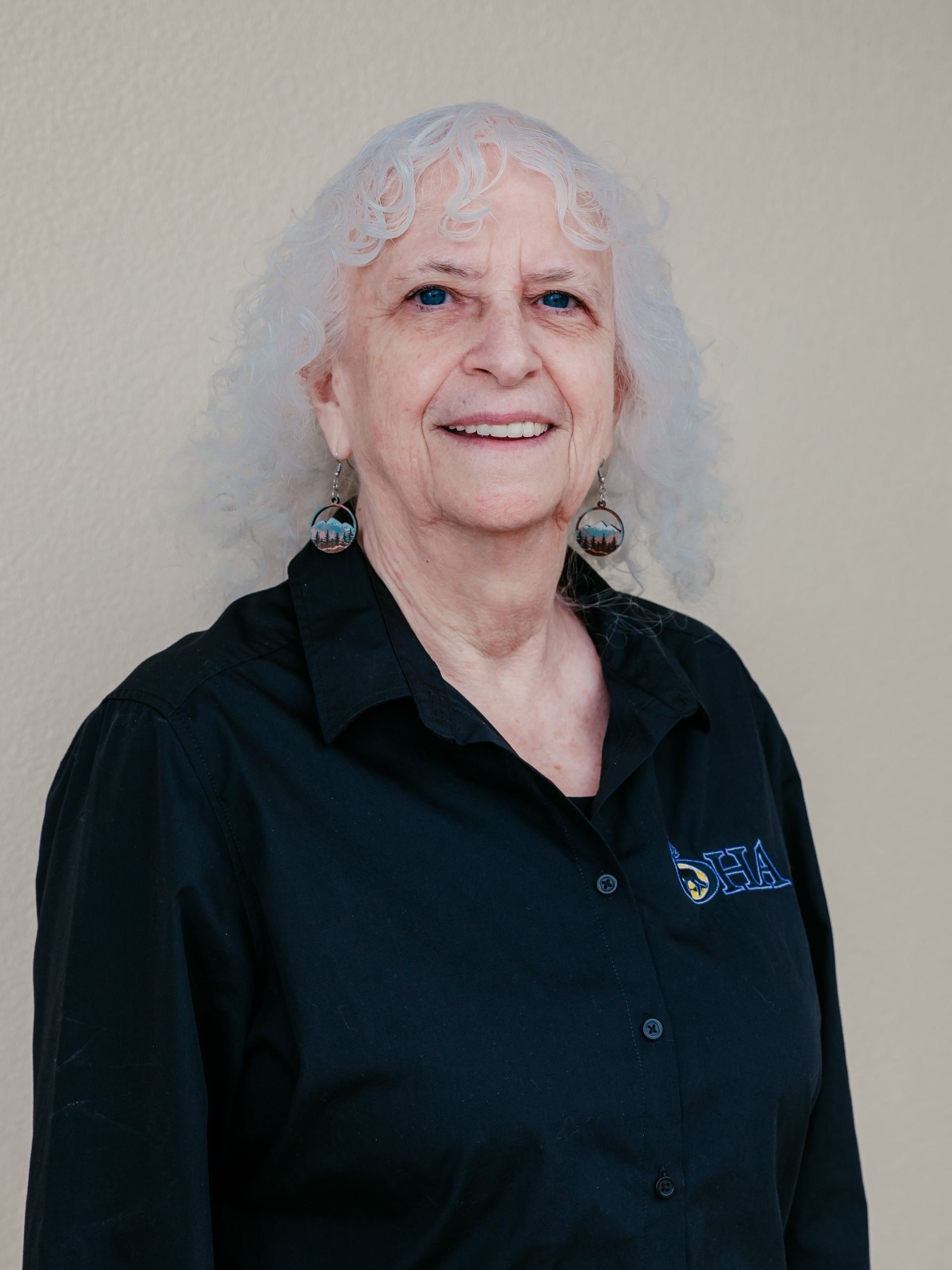 Smiling woman with curly white hair in a black shirt against a plain beige wall