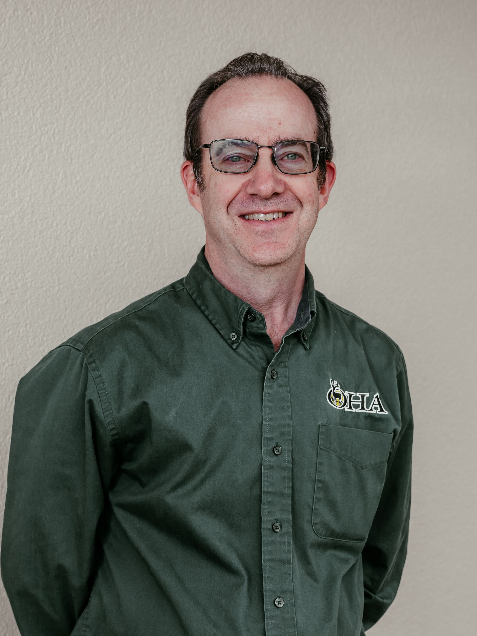 Man in a green button-up shirt and glasses, smiling against a plain wall