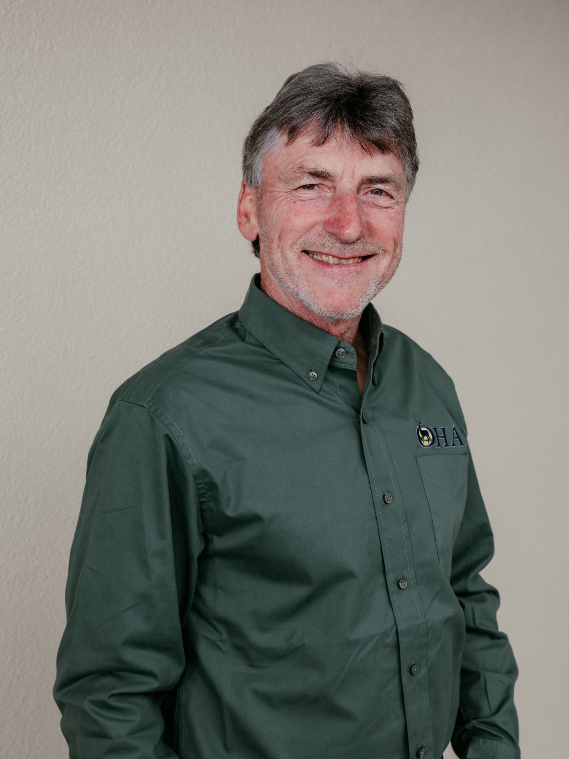 Man in a dark green button-up shirt standing against a plain light wall, smiling at the camera