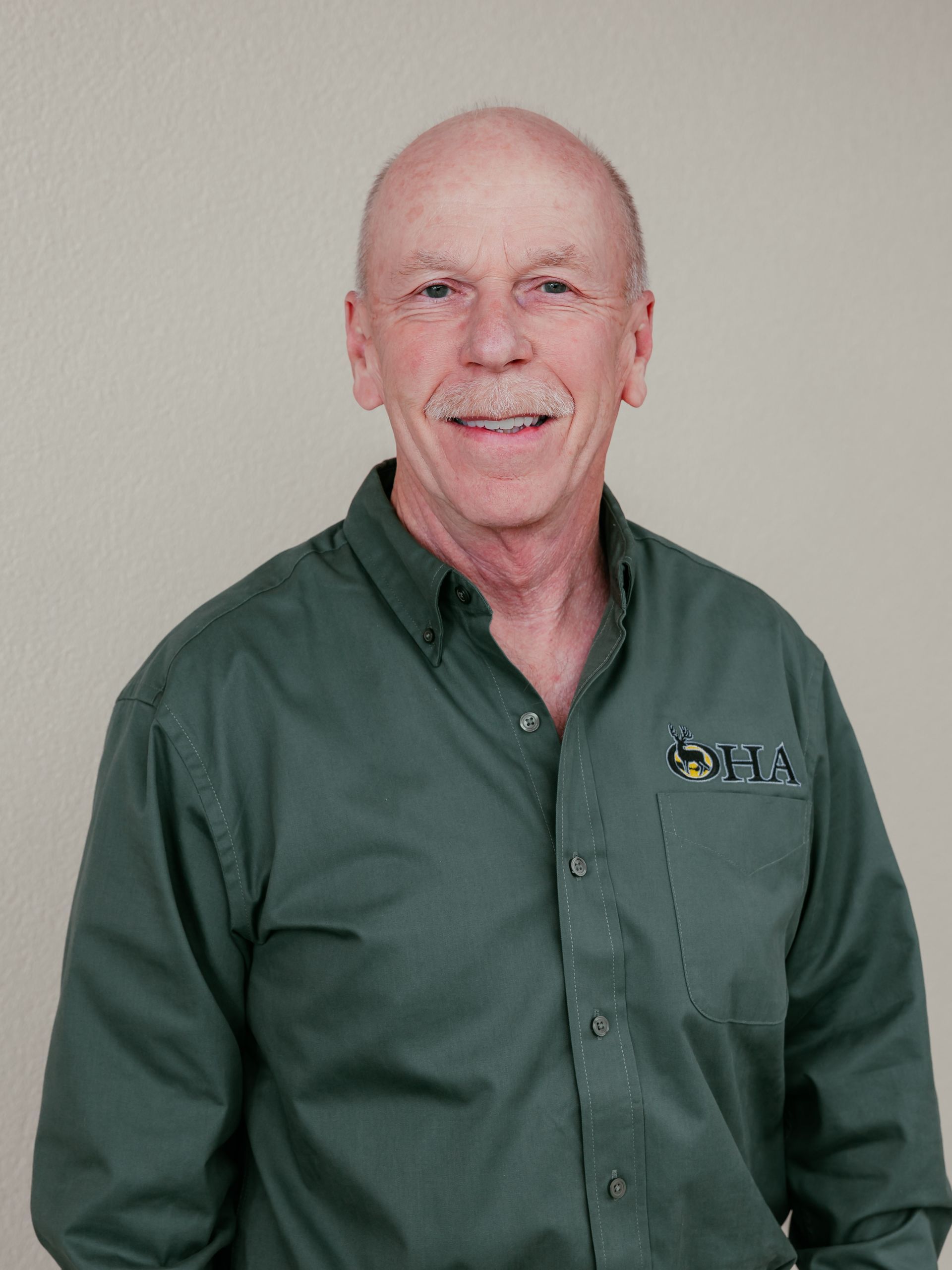 Man in a green button-up shirt standing against a plain light wall and smiling lightly