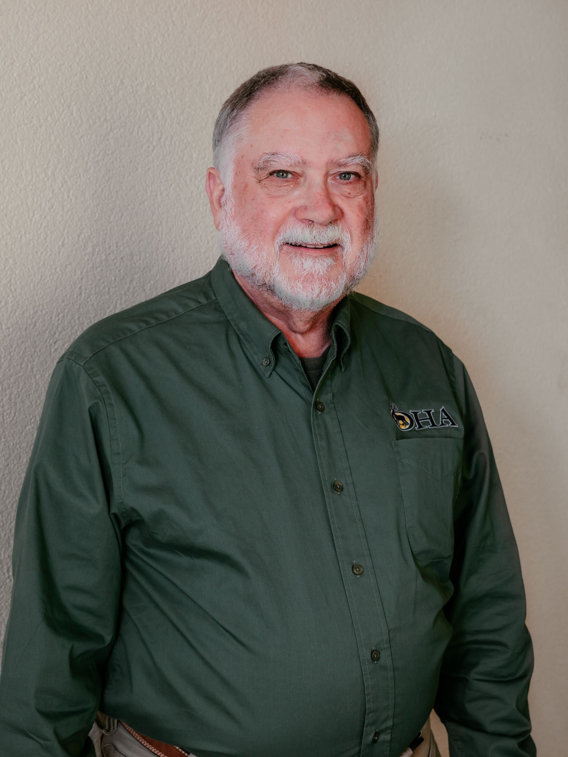 Man standing against a plain wall, wearing a dark green button-up shirt and smiling slightly.