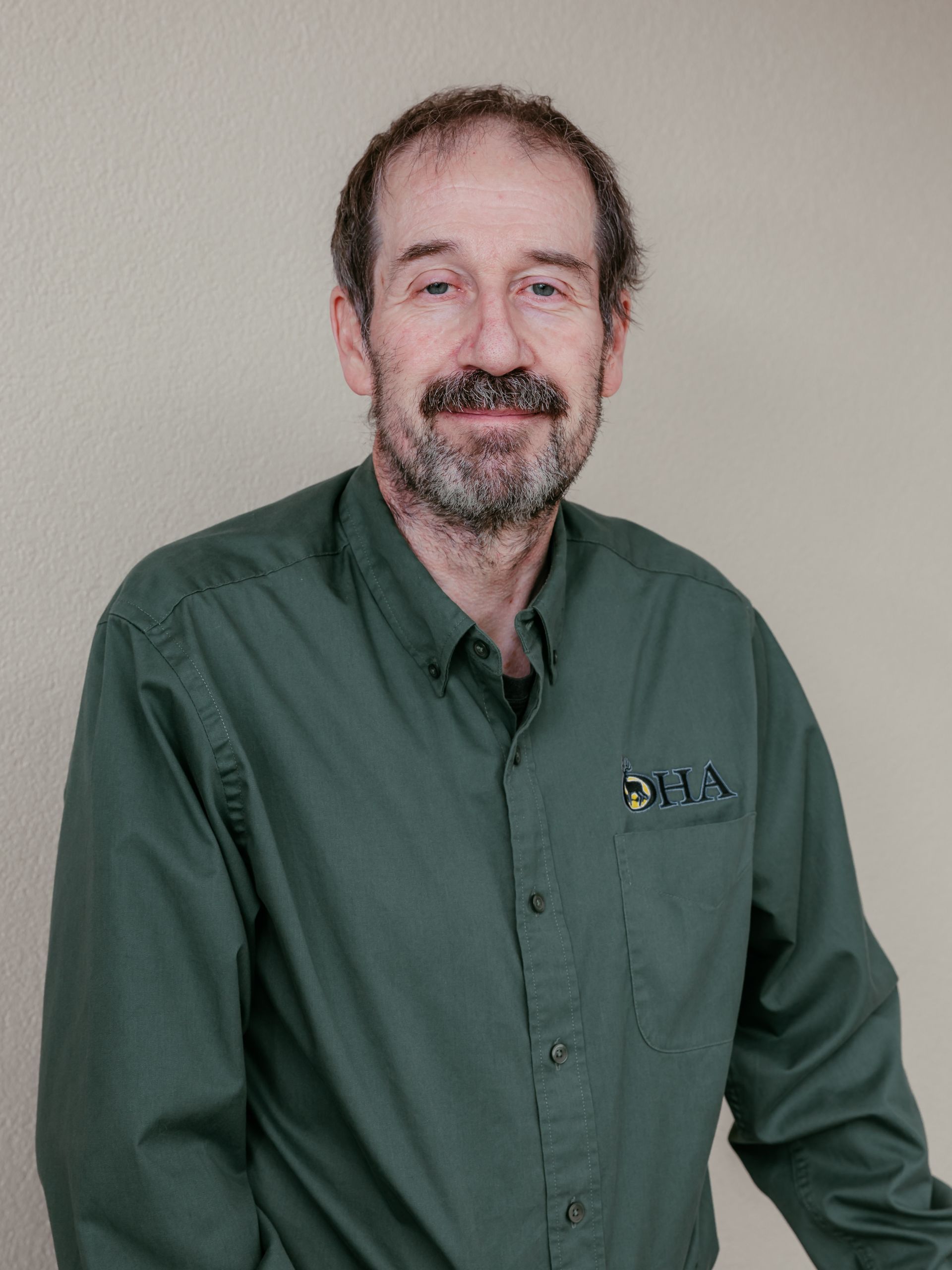 Smiling man in a green button-up shirt standing against a light wall