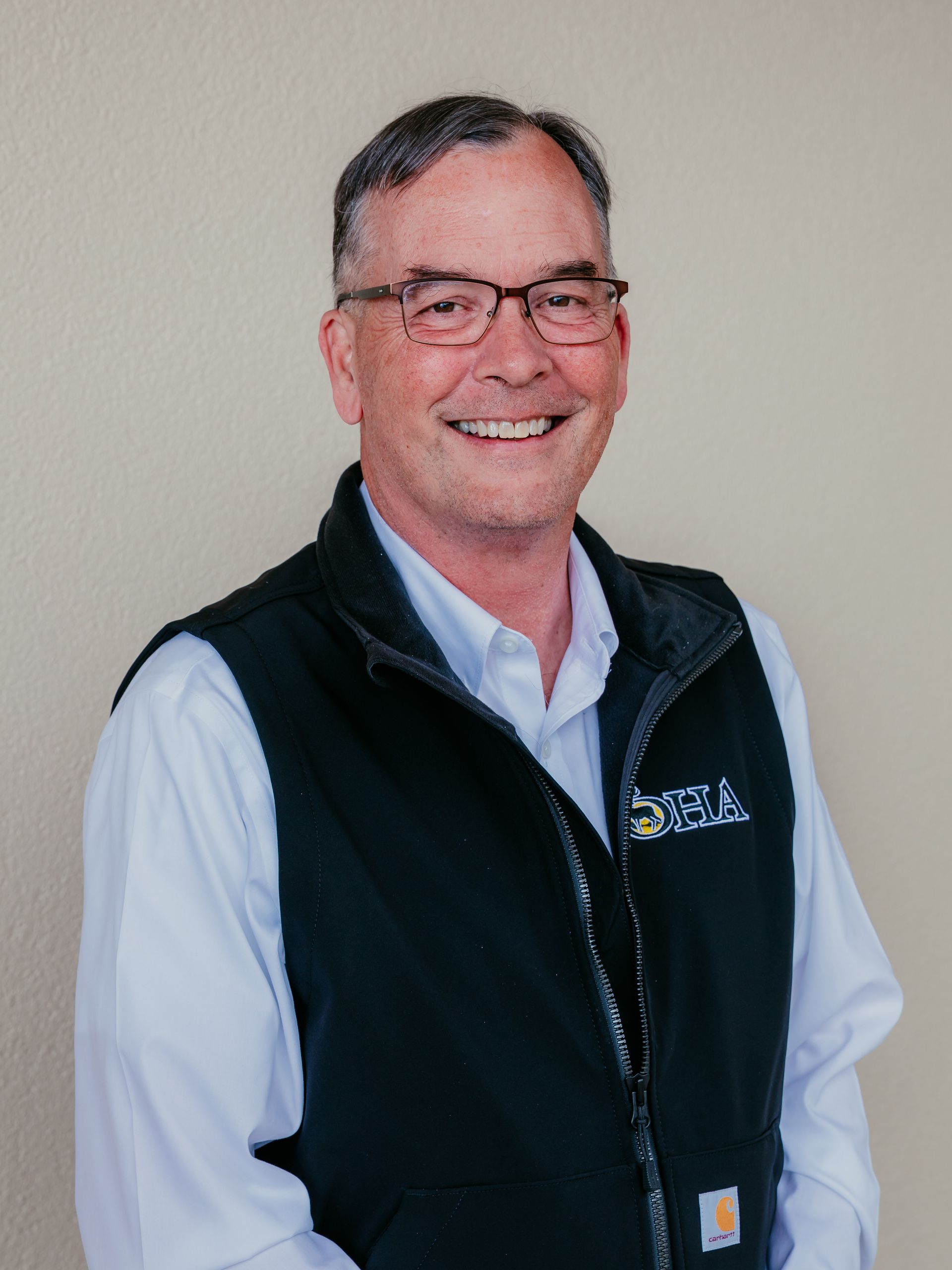 Smiling man in glasses wearing a black vest over a white shirt against a plain wall