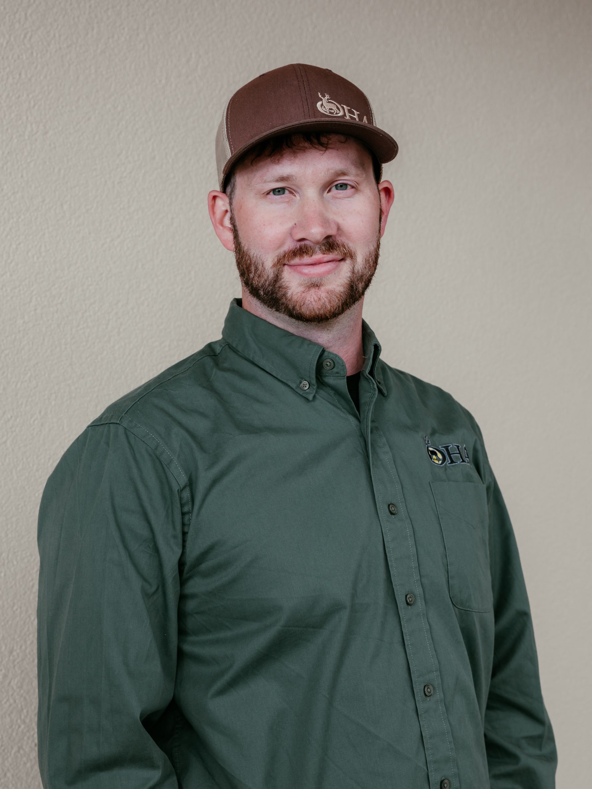 Smiling person in a brown cap and green button-up shirt standing against a beige wall