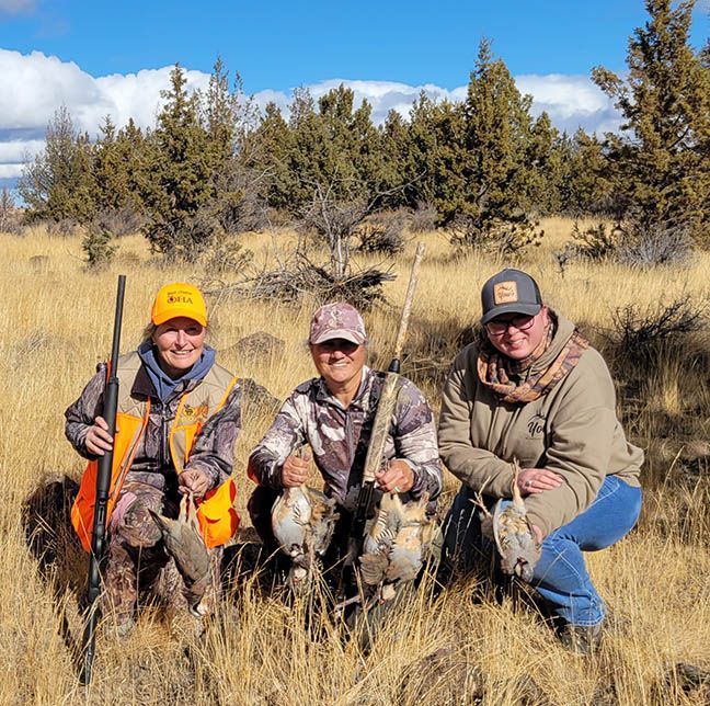 Three hunters kneeling in dry grass, each holding a bird, with rifles and a forested hillside behind them.