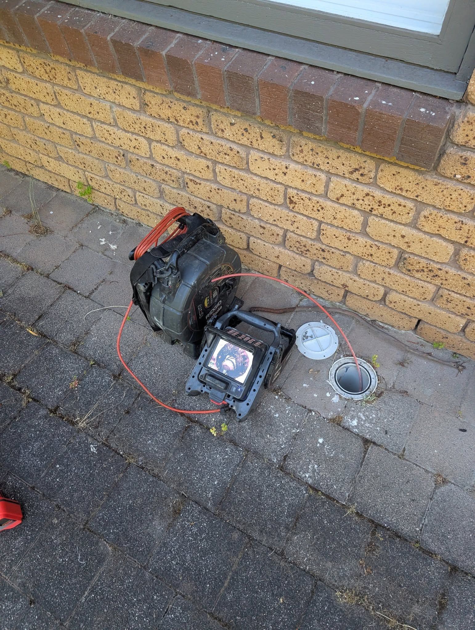 A drain inspection camera reel sits on the ground next to two open access pipes against a brick wall. — TasWide Plumbing in Kingston, TAS