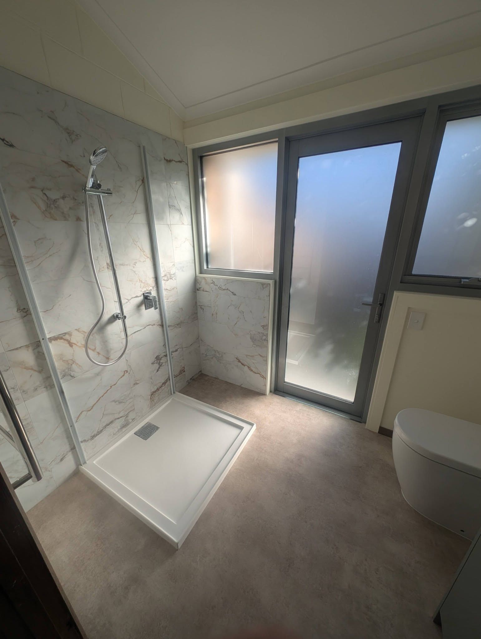 A modern bathroom with a white shower base, marble-tiled wall, handheld showerhead, and a frosted glass door. — TasWide Plumbing in Kingston, TAS