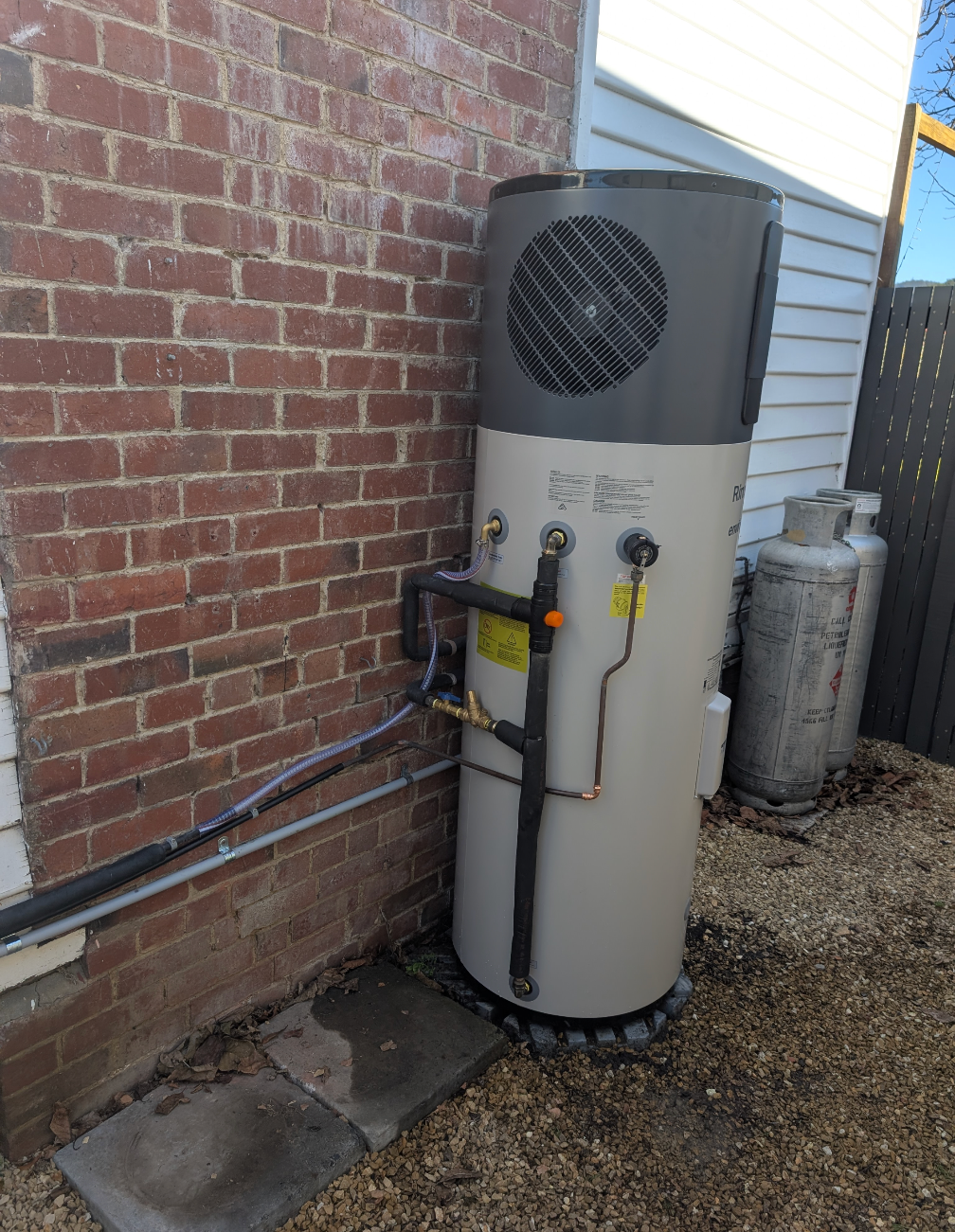 Outdoor cylindrical water heater beside a brick wall, with pipes and gravel ground. — TasWide Plumbing in Kingston, TAS